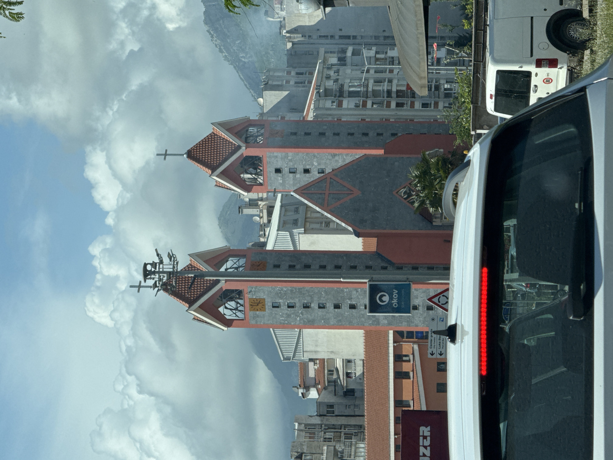The image shows the Orthodox Church of St. John Vladimir in Bar, Montenegro, viewed from a vehicle, possibly a van, on a street. The church has a distinct architectural style with gray and red roofs, topped with crosses. In the background, there are clouds and a mountain range. The surrounding area includes buildings and infrastructure typical of an urban setting. A sign with the "okov" logo is visible near the church. In the foreground, part of the vehicle's interior and rear light are visible. There is a sign indicating "Centar grada City Center" and "Luka Bar Port of Bar," suggesting the location is near the city center and the port.