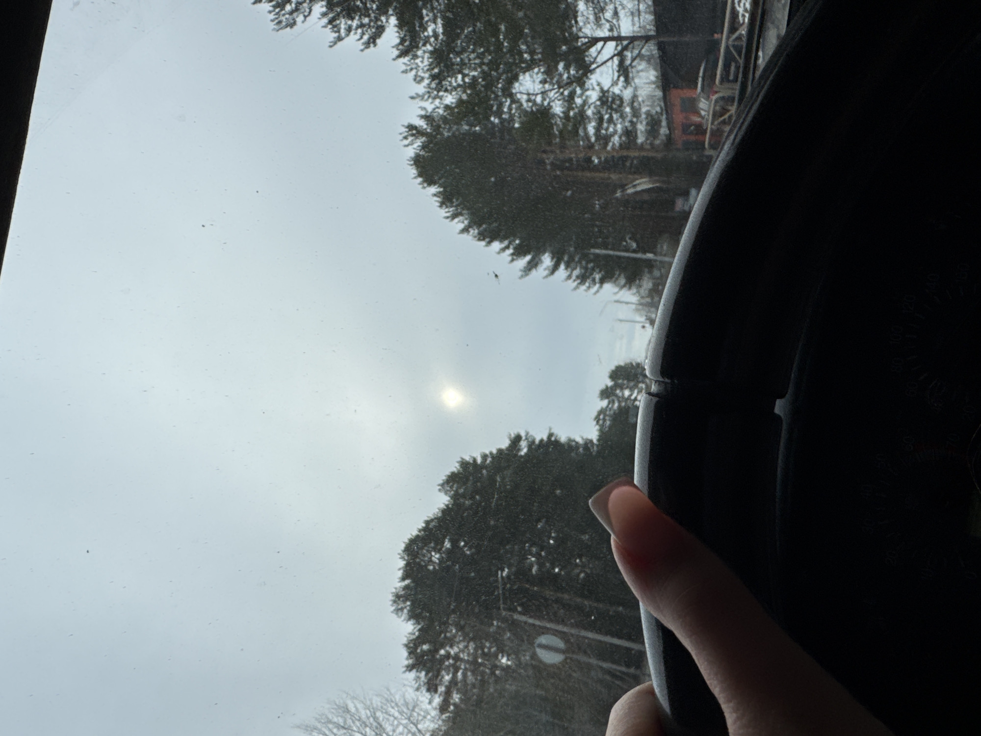 The image shows a view from inside a vehicle, looking out through the windshield. The steering wheel is visible in the foreground, held by a hand. Outside the window, trees are visible against a cloudy sky. The sun is partially obscured by the clouds. The location appears to be Chaikovsky, Russia. A road sign is partially visible, suggesting a street setting.
