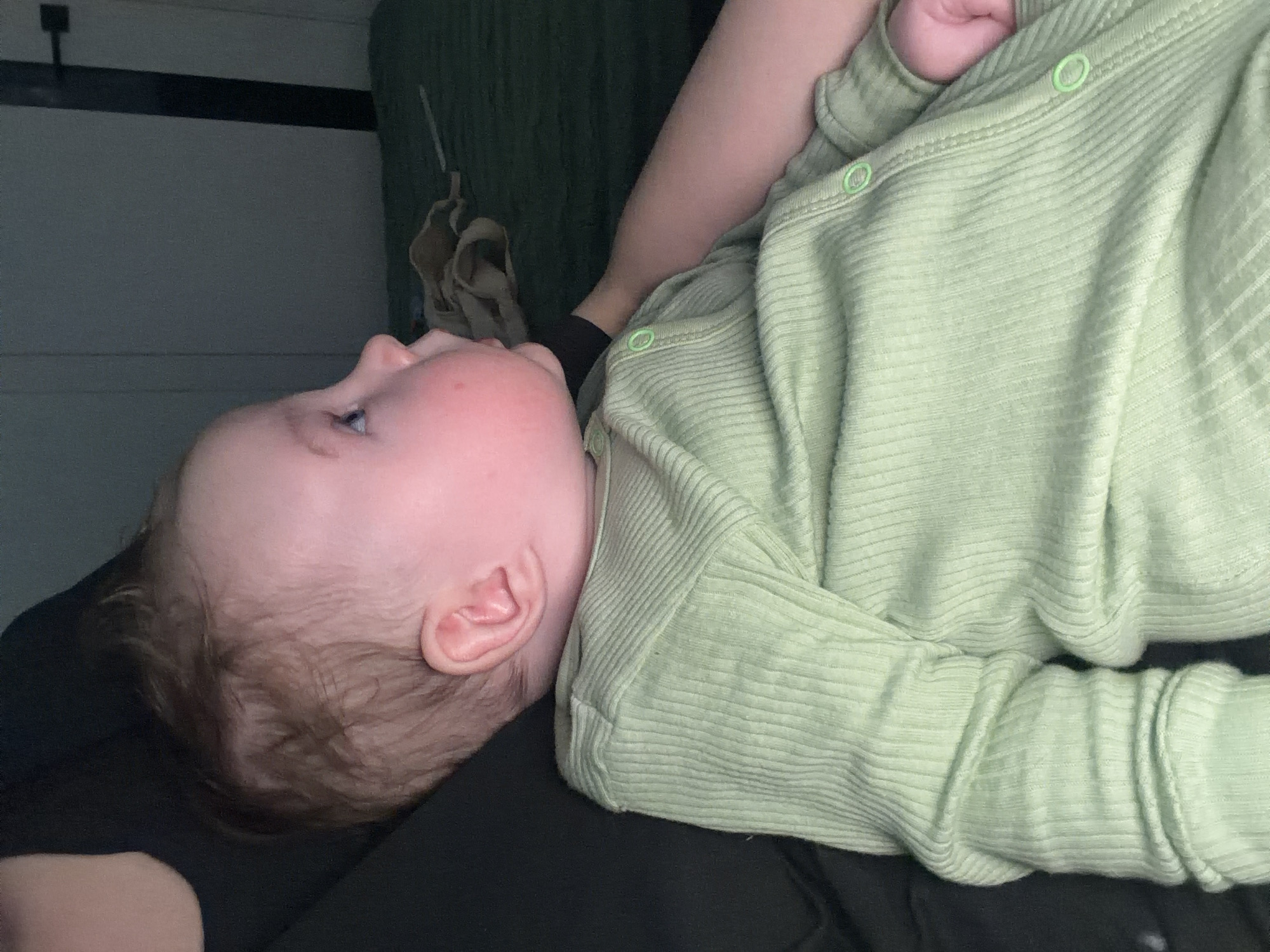 A baby wearing a light green ribbed onesie is lying on a person dressed in black clothing. The baby is positioned on their side with their head resting on the person's chest. The baby's face is visible, showing features like an ear, eye, and nose. In the background, there is a wall partially visible. Based on the prompt, this image could have been taken in the city of Chaikovsky, Russia.
