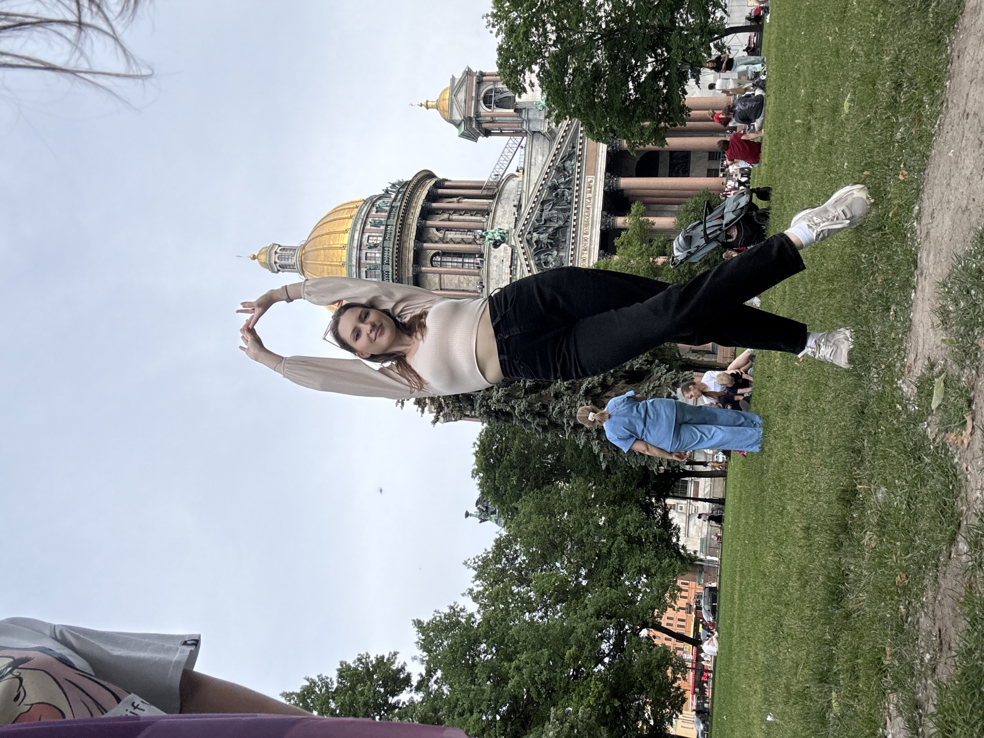 A young woman is posing with her arms raised in the air, forming a "W" shape with her hands, in front of Saint Isaac's Cathedral in Saint Petersburg, Russia. She is wearing a light-colored crop top, black pants, and white sneakers. The background features a green lawn with several people sitting and walking. A woman in blue scrubs is standing and looking away from the camera. The sky is overcast.