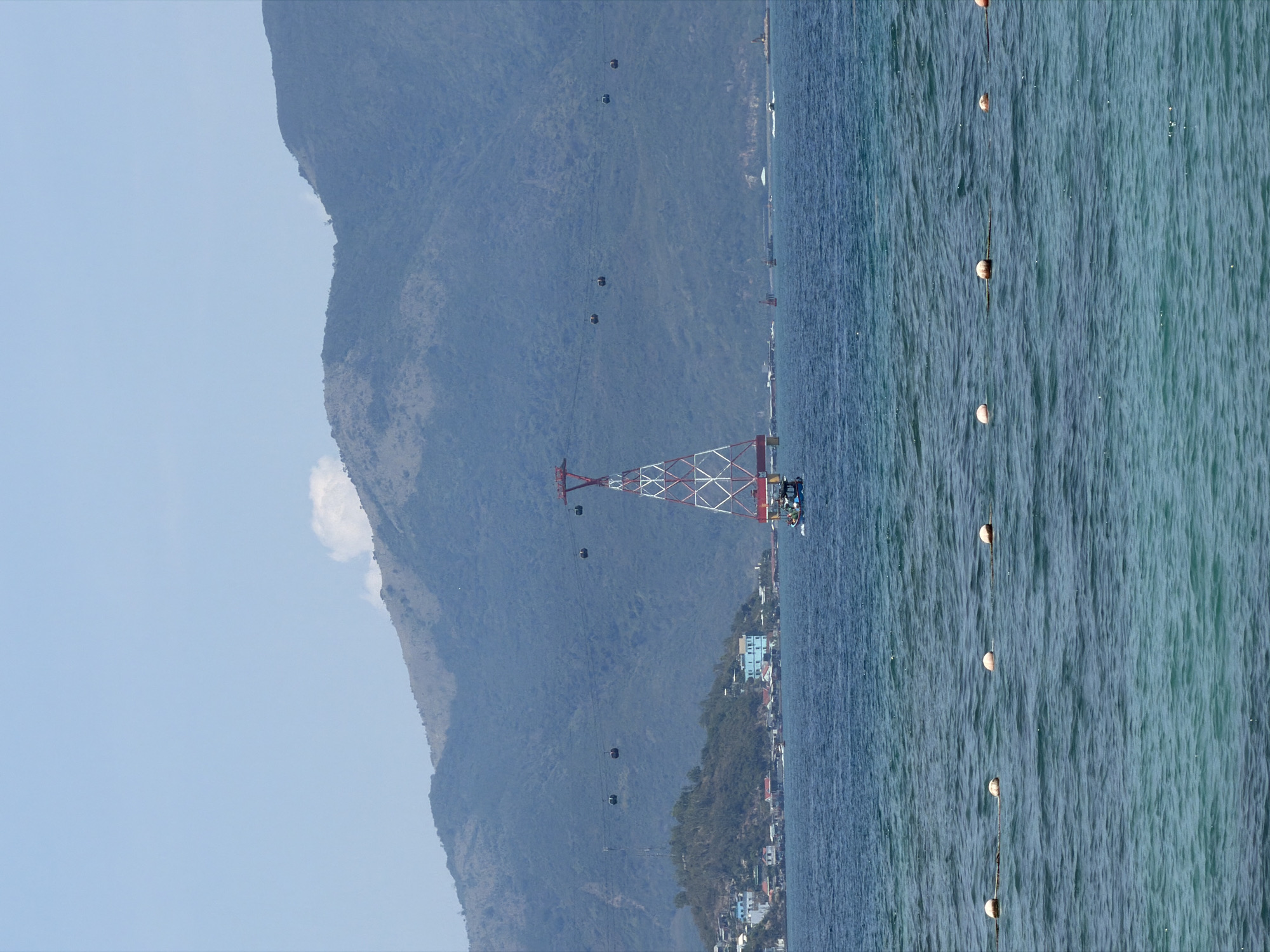 The image shows a coastal scene in Nha Trang Ward, Vietnam. A mountain occupies the left side of the frame, with a cable car line running along its slope. The cable car's support tower, painted in red and white, stands at the shoreline. Adjacent to the tower is a small cluster of buildings. The right side of the image is dominated by the ocean, delineated by a row of buoys.