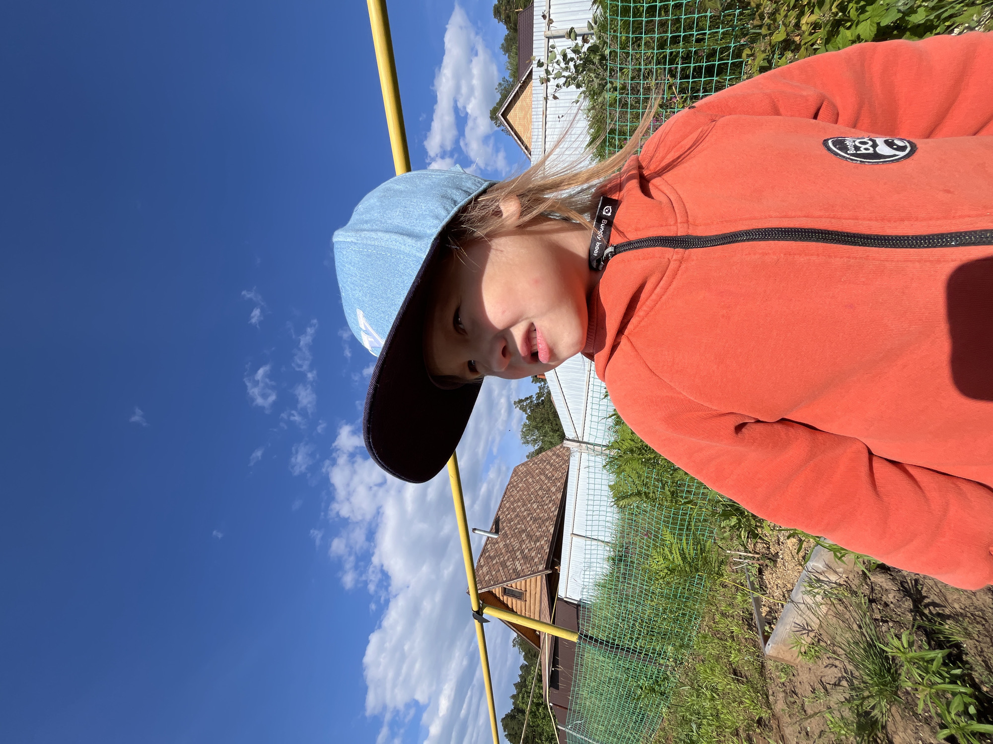 The image displays an outdoor, daytime scene in Chaikovsky, Russia, under a bright blue sky with scattered white cumulus clouds. A young child, with fair skin and light-colored hair, is positioned in the foreground, facing towards the left side of the frame with a slightly upward gaze and a focused expression. The child wears an orange hooded jacket with a black zipper closure and a circular black and white patch featuring a panda-like logo on the left chest. A light blue, denim-textured baseball cap with a dark blue brim and a white emblem is worn backwards. Sunlight illuminates the child's left side, creating shadows on the right side of the face and jacket.
In the mid-ground, a horizontal yellow metal pipe extends across the upper half of the frame. Below the pipe and behind the child, a green wire mesh fence is visible, enclosing an area with various green vegetation and a raised garden bed with exposed soil on the lower left. Further behind the fence, a white corrugated metal fence is present. The background features residential structures, including a prominent log cabin-style house with a brown shingled roof and a small attic window, visible behind the yellow pipe. Another house with a similar roof is partially visible to the right. A line of green trees and foliage marks the distant horizon.