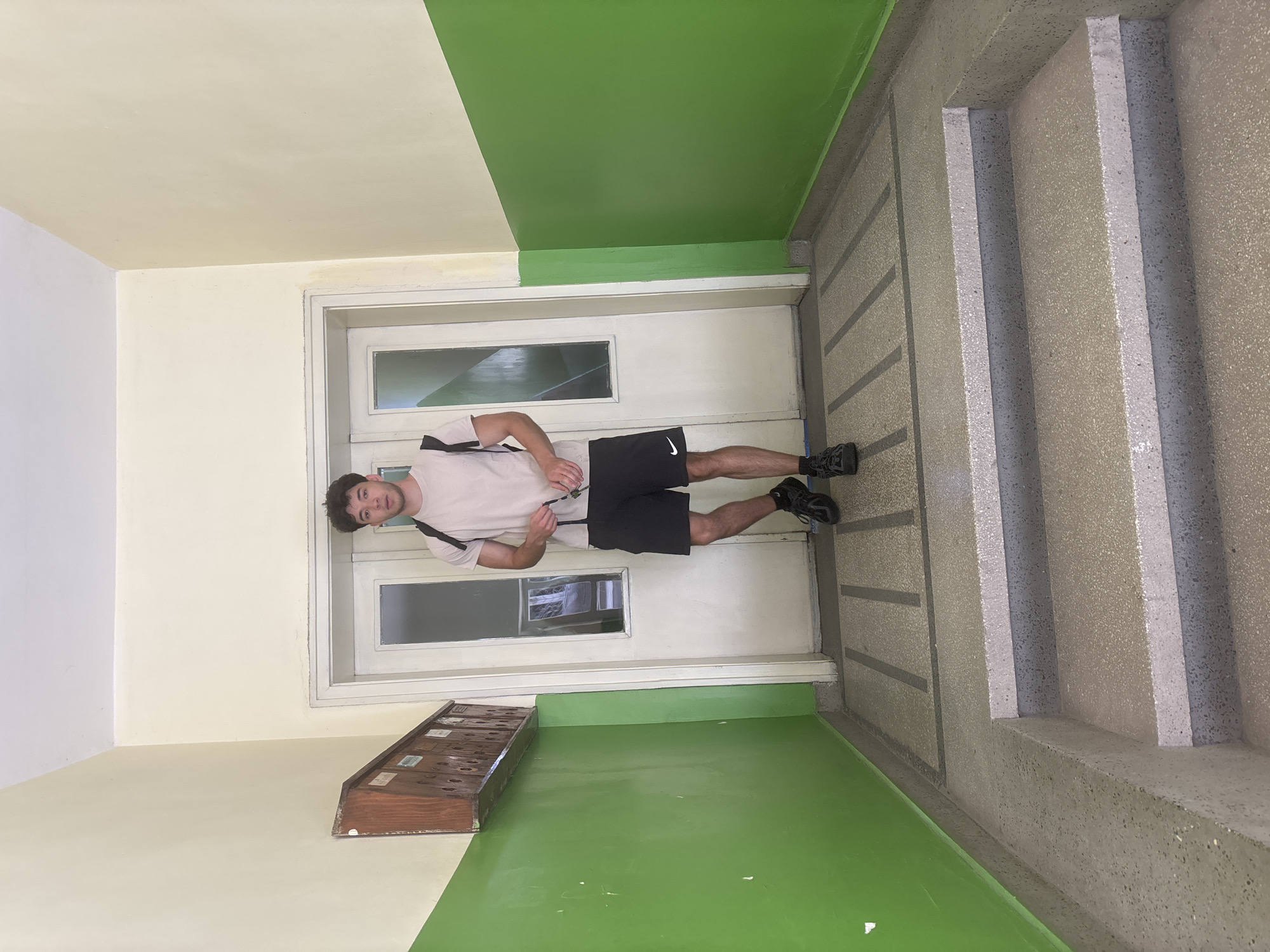 An indoor, vertically oriented photograph captures a young man seemingly standing horizontally against a wall in a hallway or stairwell, suggesting a rotated perspective. The man, with short dark curly hair, looks towards the camera with a neutral expression. He wears a light-colored t-shirt with a dark strap across his chest, black athletic shorts prominently featuring a white Nike swoosh logo on the left leg, and dark athletic shoes.
Behind the man is a white door with two rectangular glass panes. To the left of the door, a wooden mailbox unit with multiple individual compartments is affixed to the wall; small labels on these compartments are illegible. The walls are distinctly painted in two horizontal sections: a light beige/cream on the upper half and a vibrant green on the lower half. The floor and stairs are made of a light-colored material, likely terrazzo or concrete, with visible speckles and texture. A set of matching stairs with light-colored risers ascends on the right side of the frame. Horizontal textured strips are also visible on the floor near the stairs. The scene depicts a common area in what appears to be a residential building, possibly in Blagoevgrad, Bulgaria, under even indoor lighting conditions.