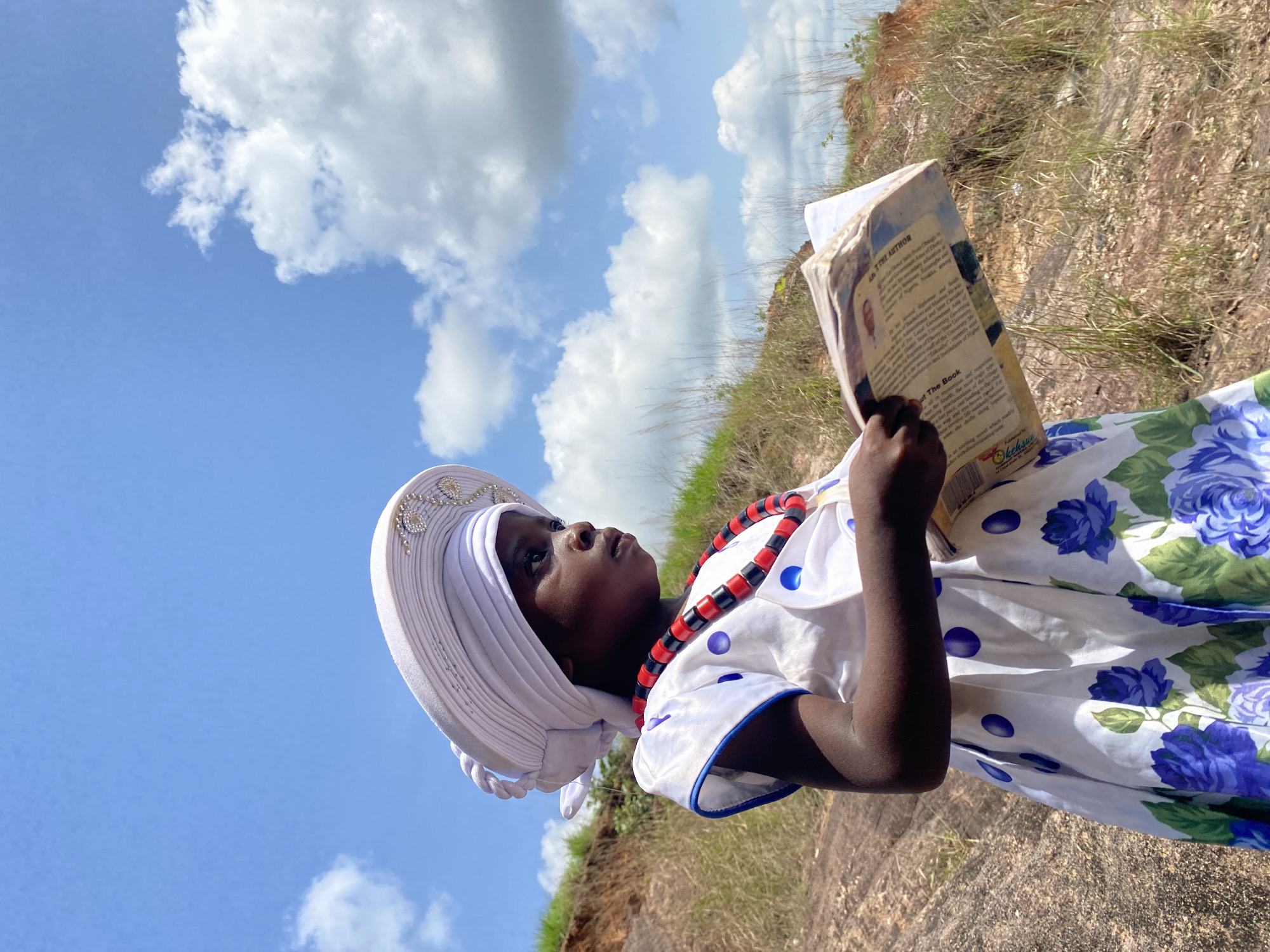 A young person is reading a book in an outdoor setting, possibly near Amaseri, Nigeria. The individual is wearing a white head wrap adorned with rhinestones, a beaded necklace of red and black, and a floral print dress featuring white, green, and blue hues. The person is holding a book titled "About the Author," with visible text and a photograph on the right page. The background consists of a blue sky with clouds and a grassy, rocky terrain.