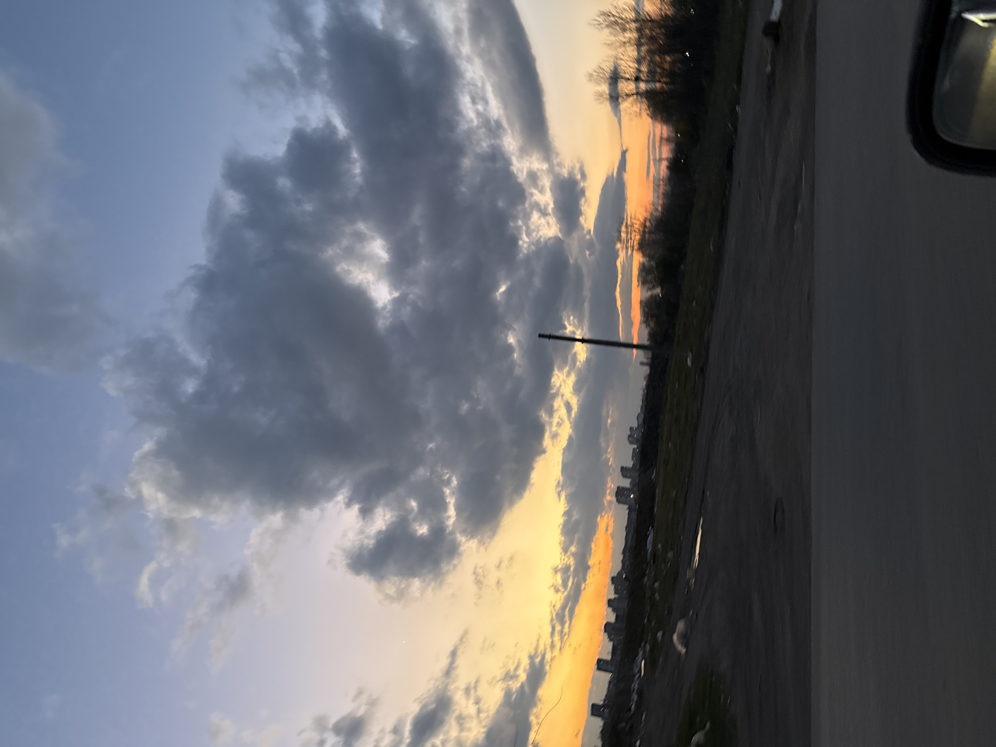 The image shows a partially cloudy sky with an orange sunset glow. The right side of the image has a section of a car body, with the side mirror visible at the top right. The left portion of the frame captures heavy, layered clouds. The ground beneath the clouds features a silhouette of a city skyline and the blurred shape of a powerline pole. It is not possible to ascertain the location in Sofia, Bulgaria from the provided information.