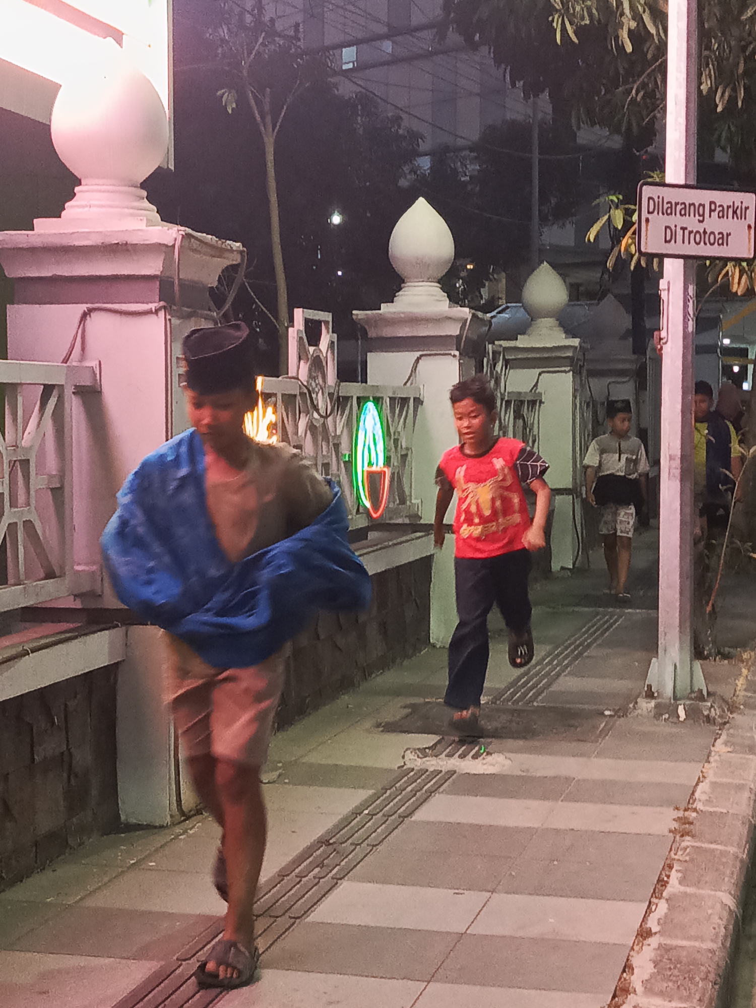 The image depicts a street scene in Surabaya, Indonesia, likely at dusk or night, with artificial lighting illuminating the area. In the foreground, a young boy wearing a black peci (a type of cap) and a tan t-shirt is walking, with a bright blue cloth draped over his shoulders. He is wearing light-colored shorts and sandals. To his right, another boy in a red t-shirt with a Spider-Man graphic and dark pants is running. In the background, other individuals can be seen walking. A sign with Indonesian text, "Dilarang Parkir Di Trotoar," which translates to "No Parking on the Sidewalk," is visible on a pole. Architectural elements such as decorative pillars and metal railings are present, along with some neon lights, suggesting a commercial or public area. The ground is paved with tiles and textured strips, indicative of a pedestrian walkway.