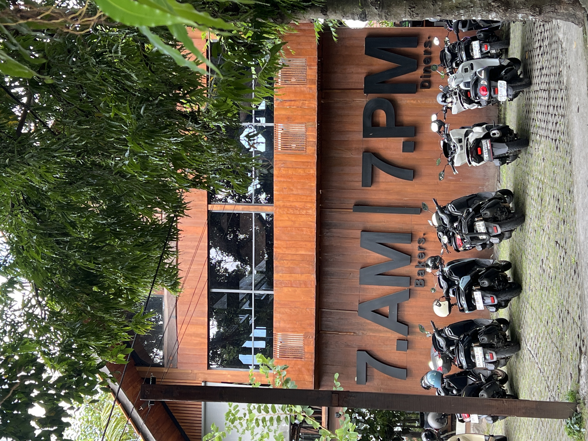 The image shows the exterior of 7.AM 7.PM Bakers. Diners. in Ubud District, Indonesia, viewed from ground level. The front facade features warm-toned vertical wooden panels. The restaurant name is prominently displayed in large, dark lettering on these panels. A vertical window section provides a glimpse into the interior.
Several motorbikes of different sizes and colors are parked in a neat row along the paved area adjacent to the building. Green foliage from trees overhangs the scene, partially obscuring the building. The scene is well-lit, suggesting it's daytime.