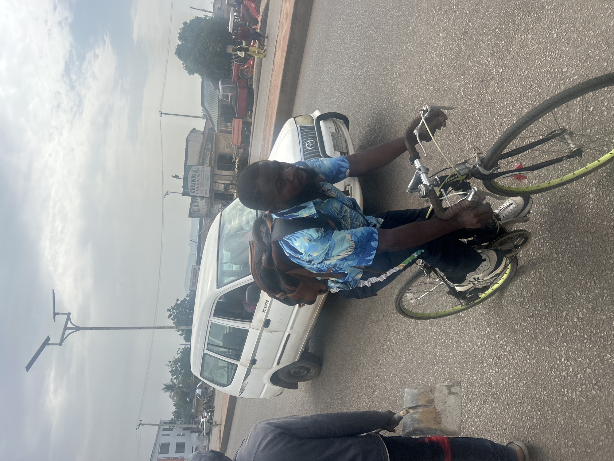 A man wearing a blue patterned shirt and a backpack rides a bicycle on a paved road in Jos, Nigeria. A white van with the license plate "JS 1100 B" is positioned close to him on the right side. Another person is visible in the lower part of the frame. The surroundings include buildings, trees, a lamppost, and other vehicles.