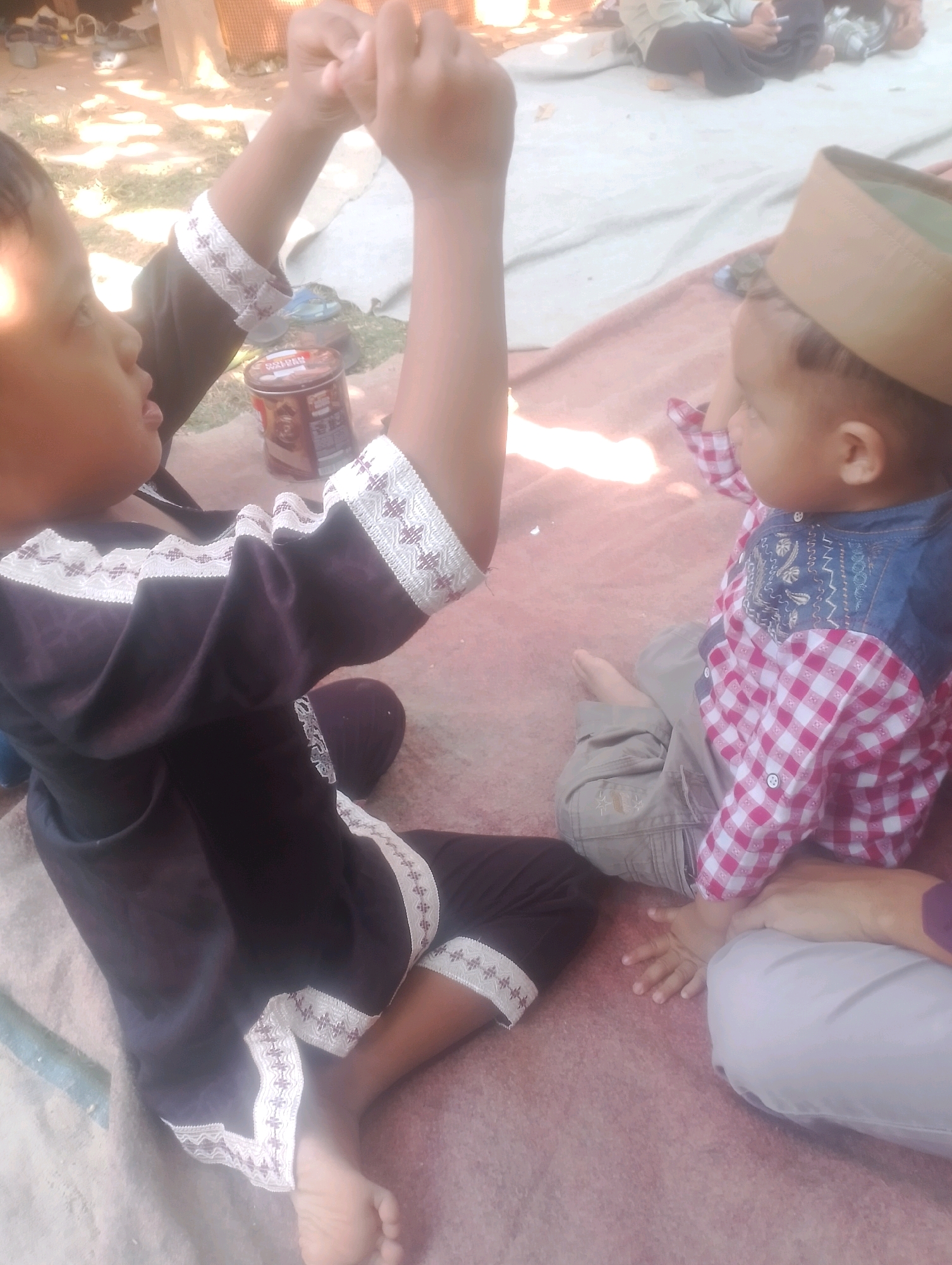 The image depicts two young children engaged in an activity outdoors on a mat in what appears to be a village setting in Kalapadua, Indonesia. On the left, a child wearing a black garment with white trim is visible from the chest up. The child is holding their hands up, clasped together, and appears to be speaking or interacting with the other child. To the right, a second child, wearing a checkered shirt and light-colored pants, is seated and wearing a tan cap. This child's hands are also visible, with one hand resting on the lap of an unseen person. The background shows a dusty ground with some scattered debris and a hint of foliage, suggesting an outdoor, possibly residential or communal area. A can, possibly of food or drink, is also present on the ground between the children.