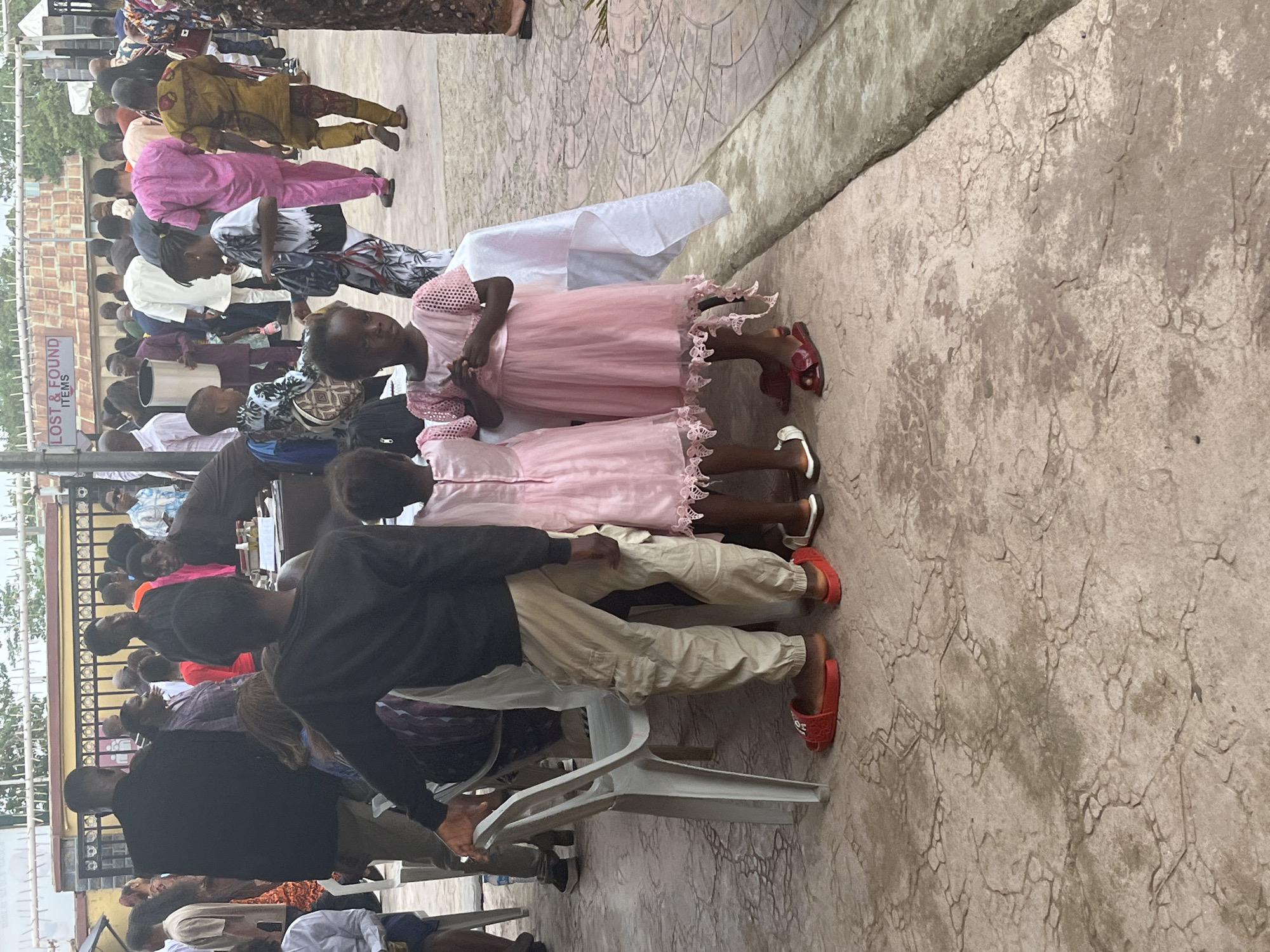 An outdoor scene shows a gathering of people in Abuja, Nigeria. In the foreground, two young girls are wearing matching pink dresses with lace detailing and different-colored sandals. A boy stands beside them, wearing a dark jacket, light-colored trousers, and red sandals. He is partially obscured by a white plastic chair. A large crowd of people of varying ages and attire sit and stand behind them. A sign labeled "LOST & FOUND ITEMS" is visible on the left side. The ground is paved with textured concrete.