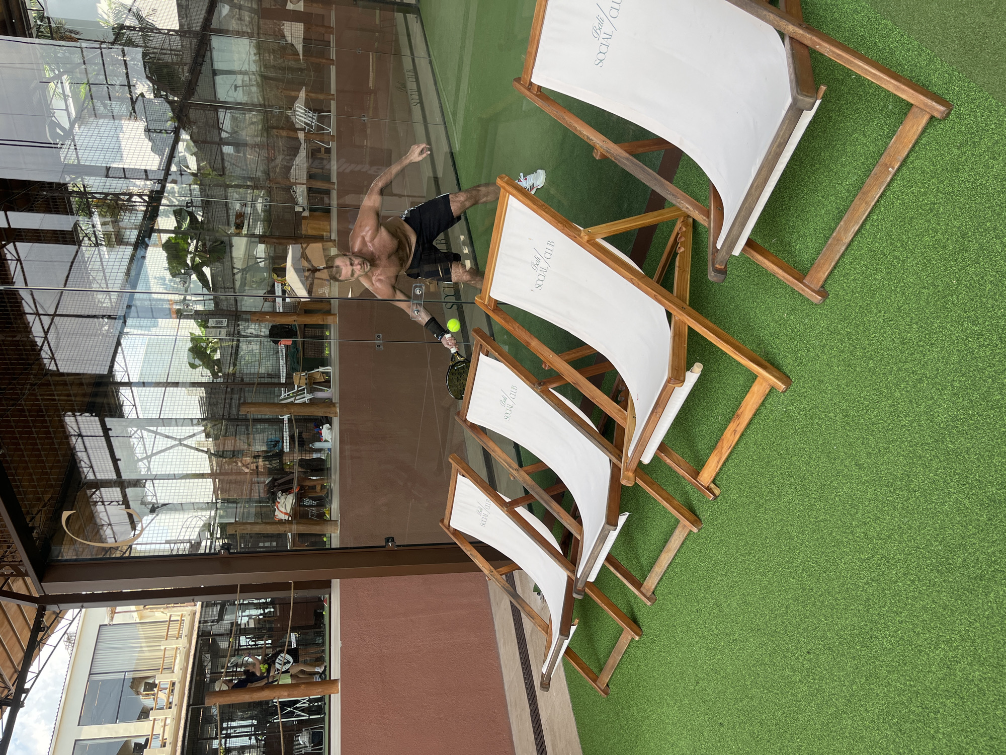 A man is photographed in North Kuta, Indonesia. He is a white, shirtless male, wearing dark shorts and white sneakers, playing padel. He has a racket in his right hand, poised to hit a yellow ball, within a padel court. The padel court has glass walls reflecting indoor plants and metal frameworks. Several white lounge chairs with wooden frames are adjacent to the court, positioned on green artificial turf. The chairs have "Bali SOCIAL CLUB" written on them. The background also features sections of a building with large windows and a brown exterior. The image is angled in a vertical orientation.