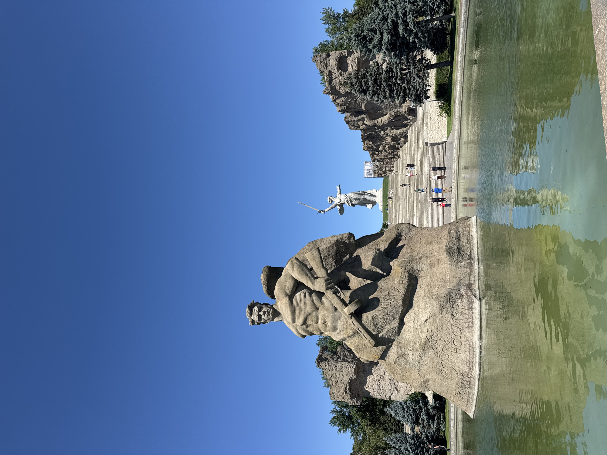 The image captures a bright, sunny day at the Mamayev Kurgan memorial complex in Volgograd, Russia. In the foreground, a massive stone sculpture of a muscular, seated male figure holding a rifle is partially submerged in a tranquil body of green water. This sculpture is inscribed with Russian text. Behind it, a long, wide set of stairs ascends a hill, flanked by rocky, textured walls. Numerous people, appearing as small figures from this distance, are visible walking up the stairs. At the top of the stairs, a towering white statue of a woman holding a sword aloft dominates the skyline. Evergreen trees are scattered throughout the scene, and the sky is a clear, vibrant blue. The overall impression is one of grandeur and solemnity, likely a tourist or memorial site.