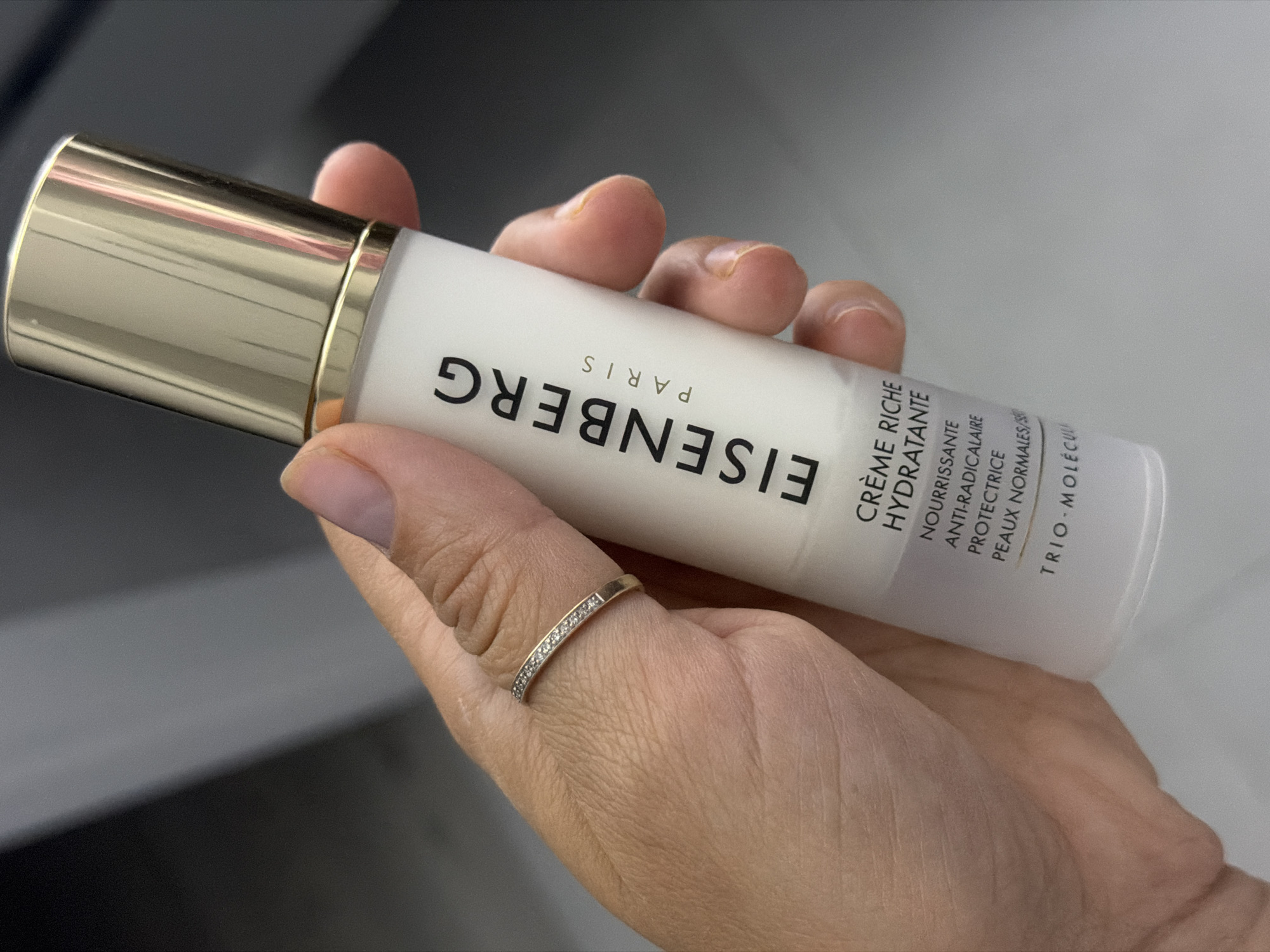 A close-up image captures a human hand holding a white and gold pump dispenser bottle of cosmetic product. The hand, exhibiting a light-to-medium skin tone, wears a thin gold band ring embedded with small, clear stones on the ring finger; the fingernails are short and natural. The bottle is vertically oriented, featuring a metallic gold cap and collar above its white body. The brand 'EISENBERG PARIS' is prominently displayed in black capital letters, with 'PARIS' stacked below 'EISENBERG'. Further down, the text identifies the product as 'CRÈME RICHE HYDRATANTE' (Rich Moisturizing Cream), followed by descriptions: 'NOURRISSANTE ANTI-RADICALAIRE PROTECTRICE' (Nourishing Anti-Free Radical Protective), intended for 'PEAUX NORMALES/SÈCHES' (Normal/Dry Skin), and employing 'TRIO - MOLÉCULAIRE' technology. The background is blurred into indistinct dark grey and black gradients, suggesting an indoor environment without specific identifiable features.