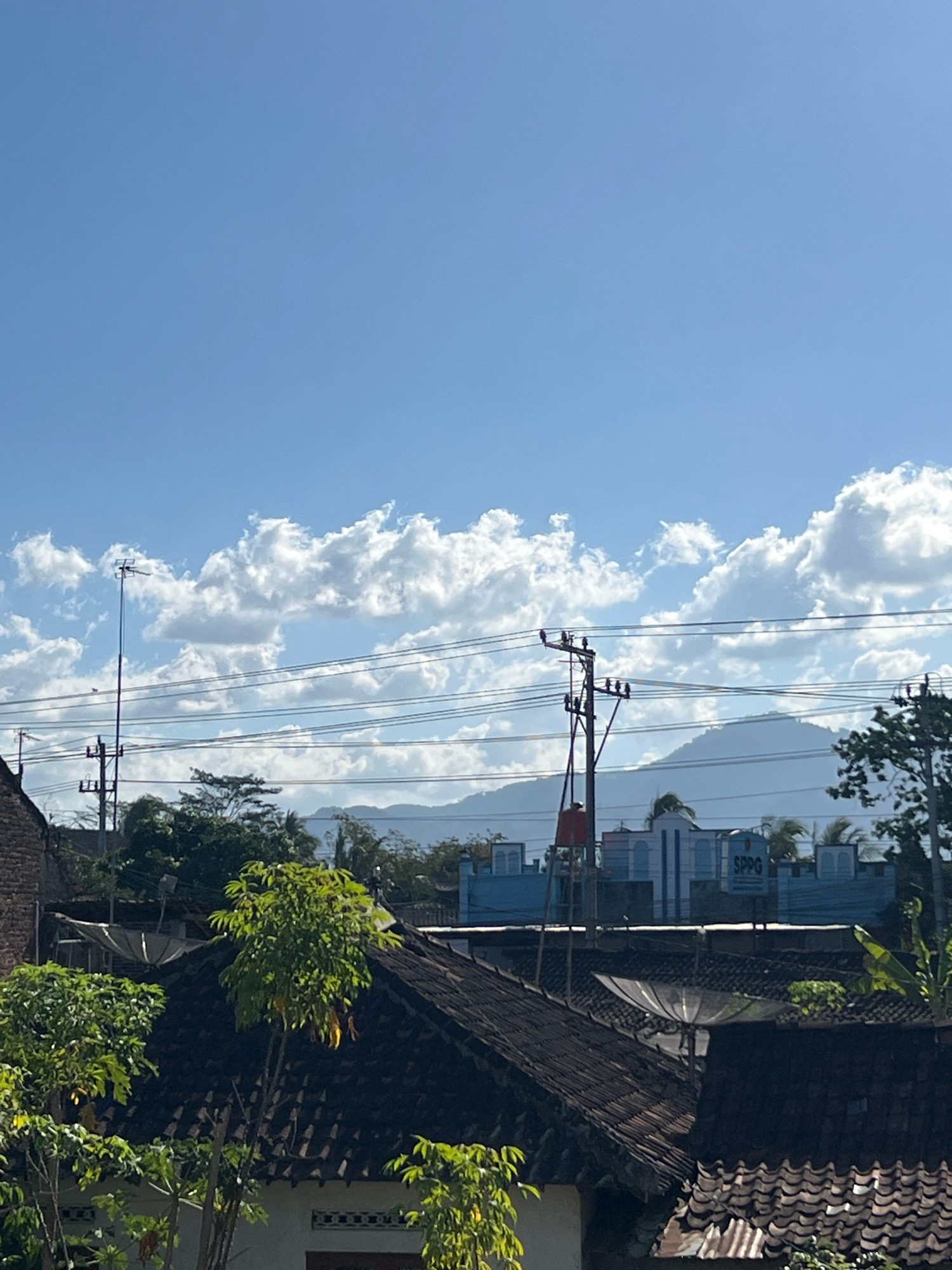 This is an eye-level, vertical, outdoor, daylight shot capturing a residential area in Payaman, Indonesia. In the foreground, dark-tiled roofs of houses are visible, with lush green trees and foliage growing around them. Satellite dishes are mounted on some of the roofs. Beyond the houses, there are utility poles with electrical wires stretching across the frame. In the mid-ground, a light blue building with white arched windows and a sign reading "SPPG" is situated. Further in the background, hazy mountains are silhouetted against a bright blue sky dotted with white, fluffy clouds. The overall impression is that of a quiet, sunny day in a rural or semi-urban Indonesian setting.