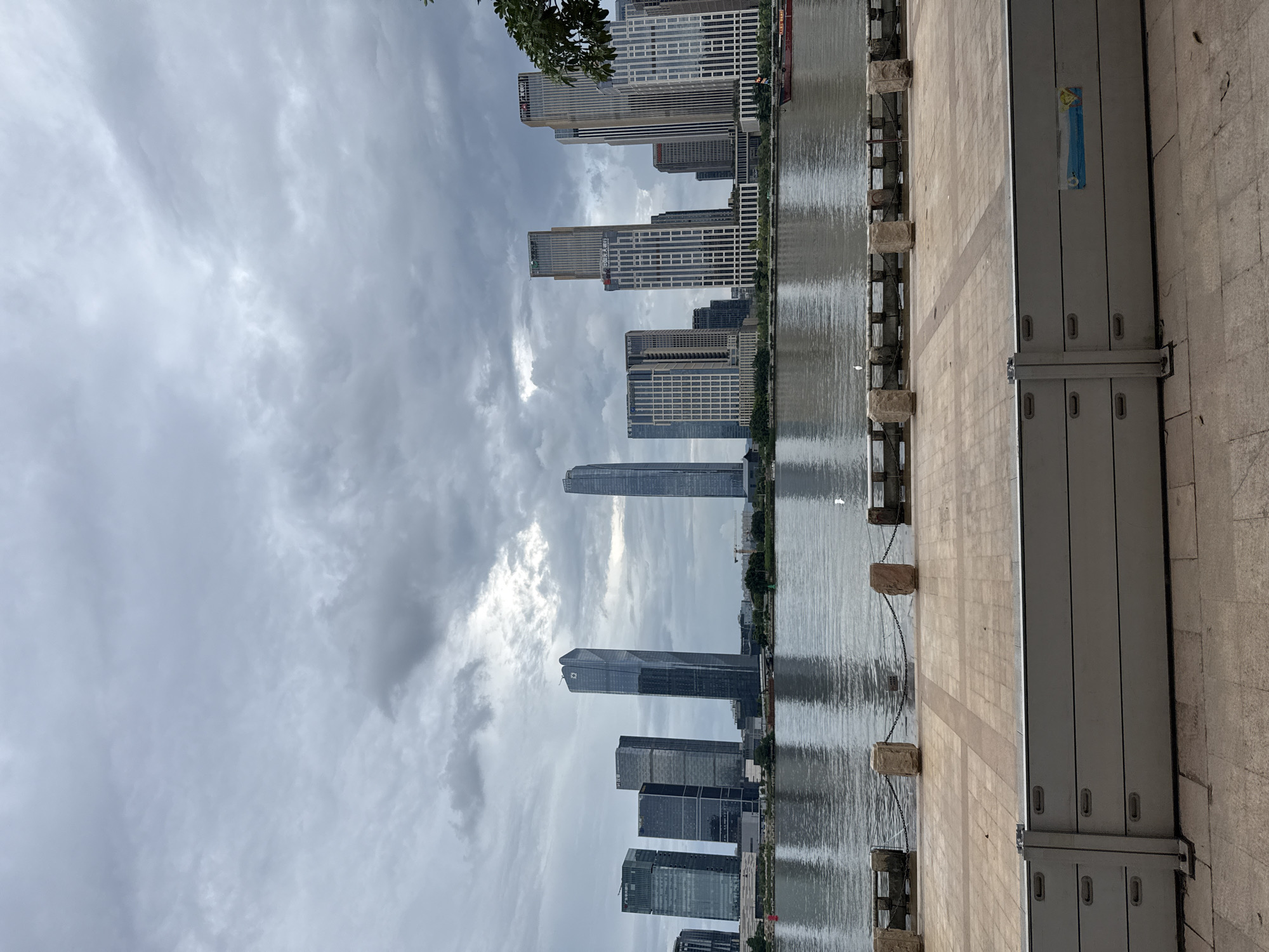 An overcast day casts a muted light over a cityscape, featuring a dense collection of modern skyscrapers on the far side of a wide river. The river's surface reflects the cloudy sky and the buildings, creating a shimmering, almost distorted image. Along the riverbank in the foreground, a paved walkway is separated from the water by a low barrier composed of large, stone posts connected by chains, and further in front, a protective flood barrier made of modular, gray panels. A few small white birds are visible on the walkway and near the water's edge. In the distance, the city skyline is dominated by tall, glass-covered buildings of varying designs. One prominent skyscraper has a unique, curved top. A red-hulled boat navigates the river. The scene suggests an urban waterfront environment within Haizhu District, China.