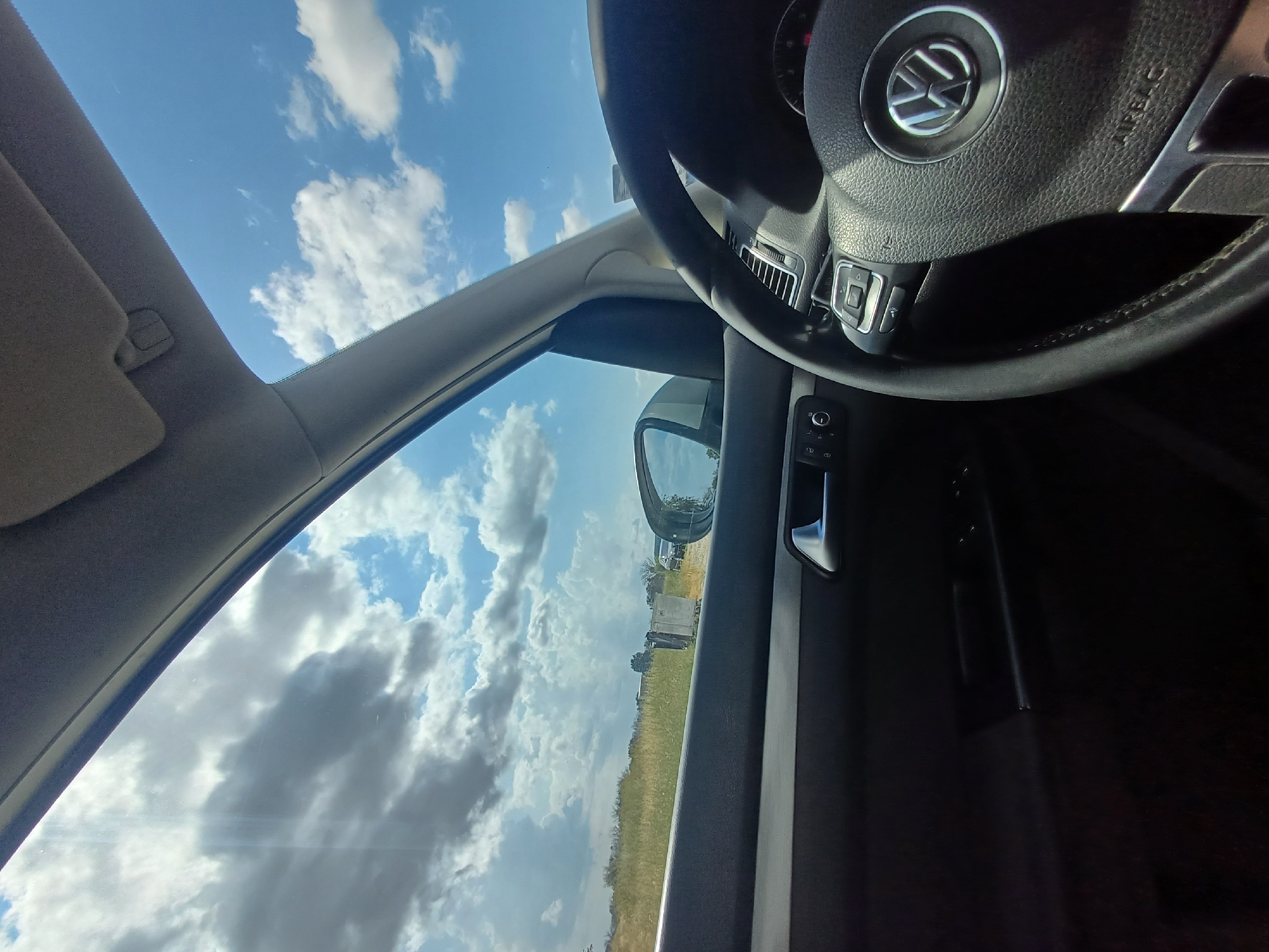 The image captures the interior of a car, looking out through the windshield and side window towards a rural landscape under a partly cloudy sky. The driver's side steering wheel, a Volkswagen emblem, and part of the dashboard are visible in the foreground. Outside, a grassy field stretches to the horizon, with some scattered trees and what appear to be structures or vehicles in the distance. The sky is a vibrant blue with fluffy white and gray clouds. The scene is set in Aksakovo, Bulgaria. There are no people visible in the image.