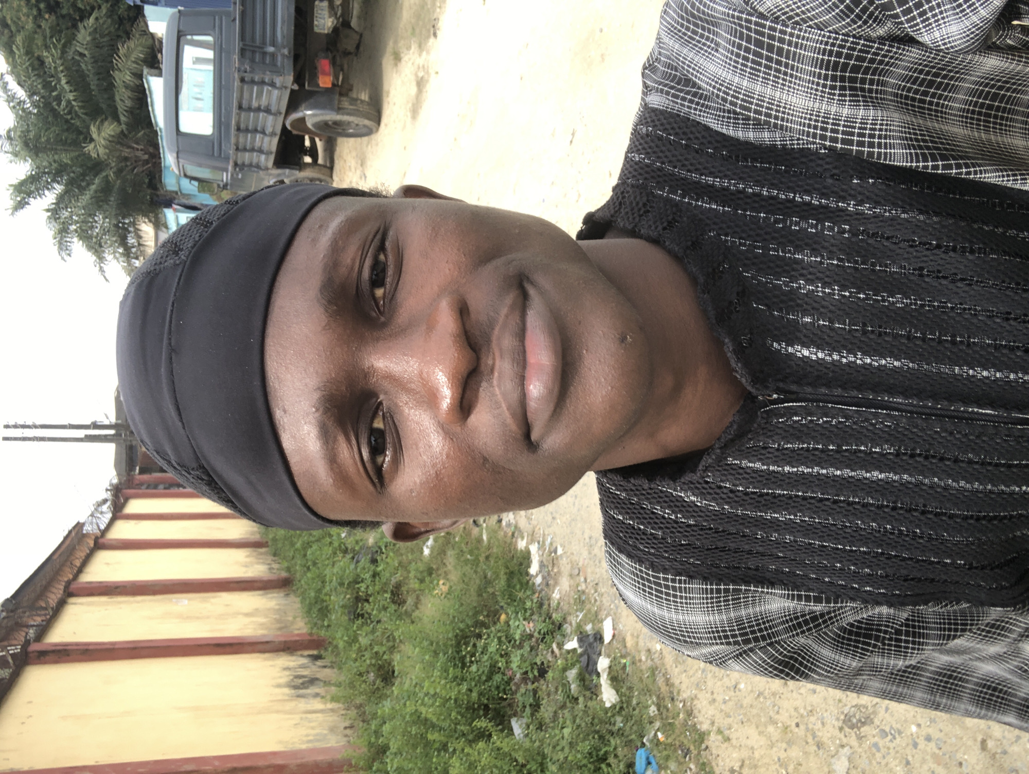 A young man with dark skin and short black hair, wearing a black skull cap and a black and white plaid shirt with black mesh panels, is standing outside. The man is looking directly at the camera and smiling slightly. The background of the image shows a yellow wall with red vertical posts, lush green bushes, a dirt road, and a dark gray pickup truck. A small section of a palm tree is visible on the right side of the image. The scene appears to be in a semi-urban area, possibly a residential street or an industrial zone, in Ado, Nigeria.