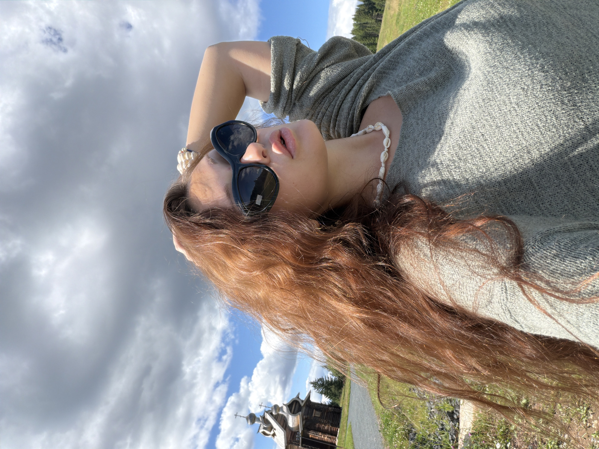 In a medium shot, a young woman with long, auburn hair stands outdoors. She is wearing a light gray, textured sweater and a necklace made of small white shells. She has on large, dark sunglasses and is posing with her right arm behind her head. Her lips are slightly parted, and she is looking upwards towards the sky. The background features a blue sky with scattered white clouds. To the left, a rustic wooden church with multiple domes and crosses is visible, situated on a gentle grassy slope. A gravel path leads towards the church, and pine trees dot the landscape beyond. The overall setting suggests a rural or historical site.