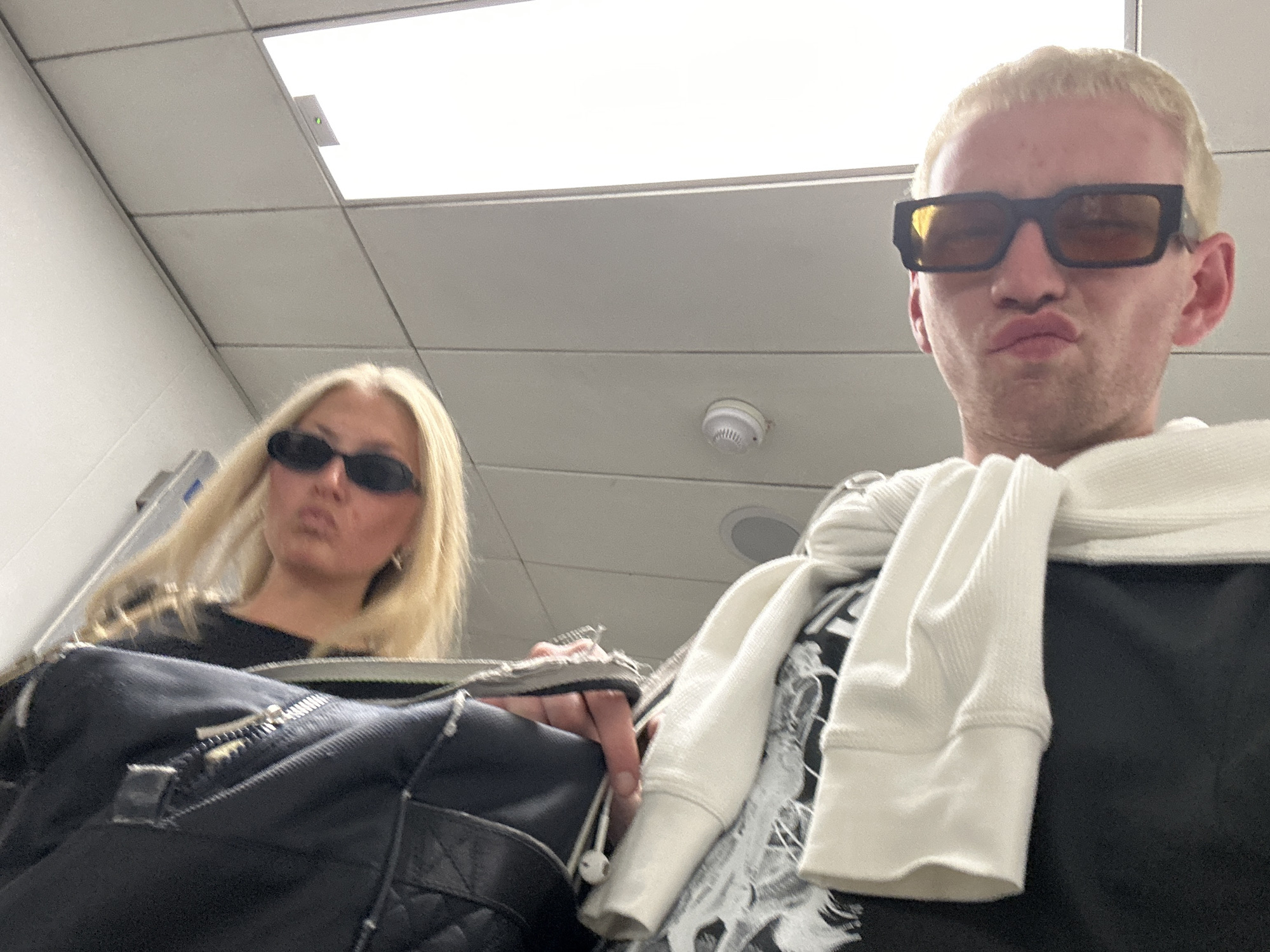 This low-angle indoor selfie features two individuals, both with blonde hair and sunglasses, making puckered lip expressions. In the foreground, on the right, a person with short bleached blonde hair wears large, dark rectangular sunglasses with amber lenses. They have a white long-sleeved top draped over their shoulders, over a dark garment. In the background, on the left, a person with long blonde hair wears small, black oval sunglasses and holds a black bag with a quilted texture and a visible zipper. White wired earphones are visible near their hand.
The setting appears to be a modern indoor space, likely a commercial or public building, indicated by the white suspended ceiling with modular tiles, bright rectangular fluorescent light panels, and a visible circular smoke detector. The expressions of both subjects convey a playful or humorous mood, possibly posing for a casual photo while waiting. The image captures a moment of lighthearted interaction within an institutional environment. No visible text is present.