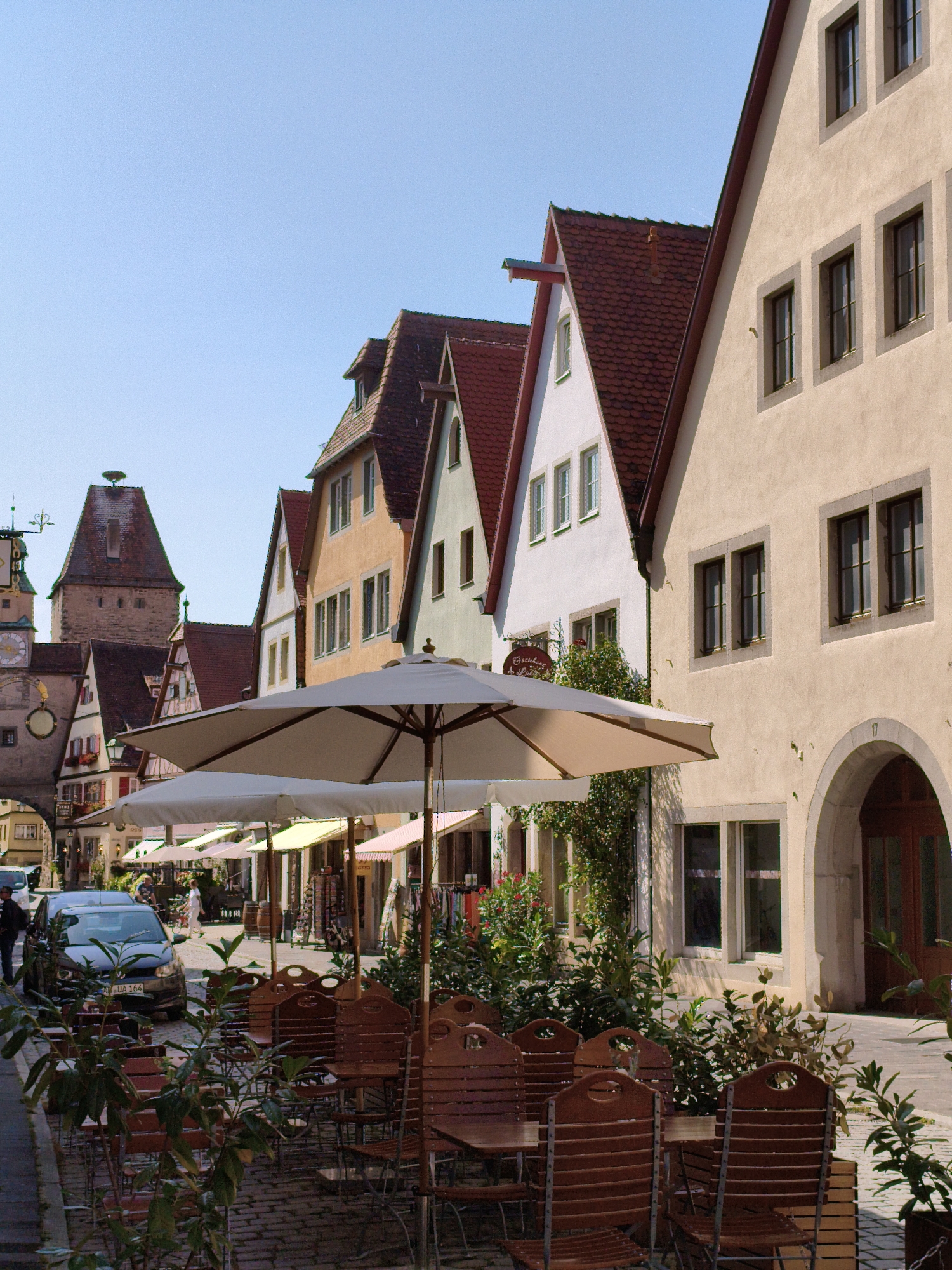 This is a photograph taken in the city of Rothenburg ob der Tauber, Germany, likely on a sunny day with clear skies. The image captures a street scene featuring traditional Bavarian architecture and an outdoor seating area for a cafe or restaurant.
In the foreground, several outdoor dining tables and chairs are arranged on a cobblestone street. The chairs are made of dark wood with slatted backs. Two large, cream-colored umbrellas provide shade over the tables. Green foliage and plants are visible around the seating area, adding a touch of nature to the scene.
Beyond the seating area, the street continues, lined with a row of historic buildings. These buildings exhibit characteristic timber-framed elements and pitched red-tiled roofs. The facades are painted in various warm colors, including cream, orange, and light green. Windows are visible on each building, some with multiple panes.
Further down the street, a prominent tower with a clock face stands tall, a common feature of Rothenburg ob der Tauber's medieval cityscape. A stork's nest is visible on top of this tower. Cars are parked along the street, and a few people are seen walking or standing, suggesting a public area. One dark-colored car with a German license plate is parked in the middle ground.
The overall impression is one of a picturesque, historic European town that is open for business and tourism, with its charming architecture and inviting outdoor spaces.