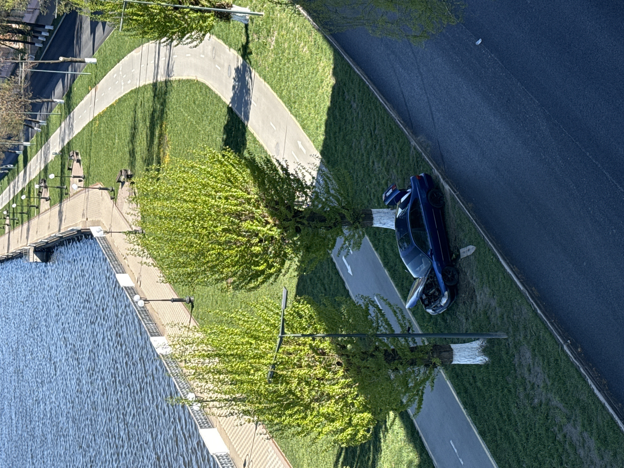 An aerial view shows a blue car on the grass between a road and a bike path. The car's hood is open. Next to the bike path is a lake. The location appears to be near Votkinsk, Russia.