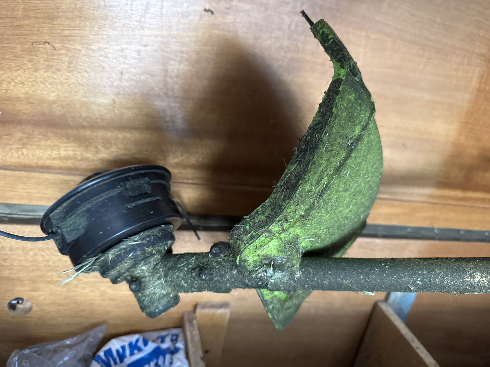 The media file displays a close-up view of a string trimmer head, covered in dried grass and dirt. The trimmer head is black with a visible spool of string. A green guard, also caked with debris, is attached to the head. The trimmer is resting horizontally on a wooden surface, likely a shelf or workbench, in what appears to be an indoor storage area. The background consists of light brown wooden panels with visible grain. There are hints of other objects in the lower portion of the image, including what looks like plastic packaging and a portion of a blue and white item with text.
The image is focused on the trimmer head and does not show any people or a specific event taking place. The lighting suggests an indoor setting, possibly in a garage or shed, and the time of day is indeterminate. There are no weather-related cues visible. The only discernible text is on the blue and white item in the lower part of the frame, which appears to be a portion of "MYKA" in Cyrillic letters. The overall impression is that of a used piece of gardening equipment stored away.