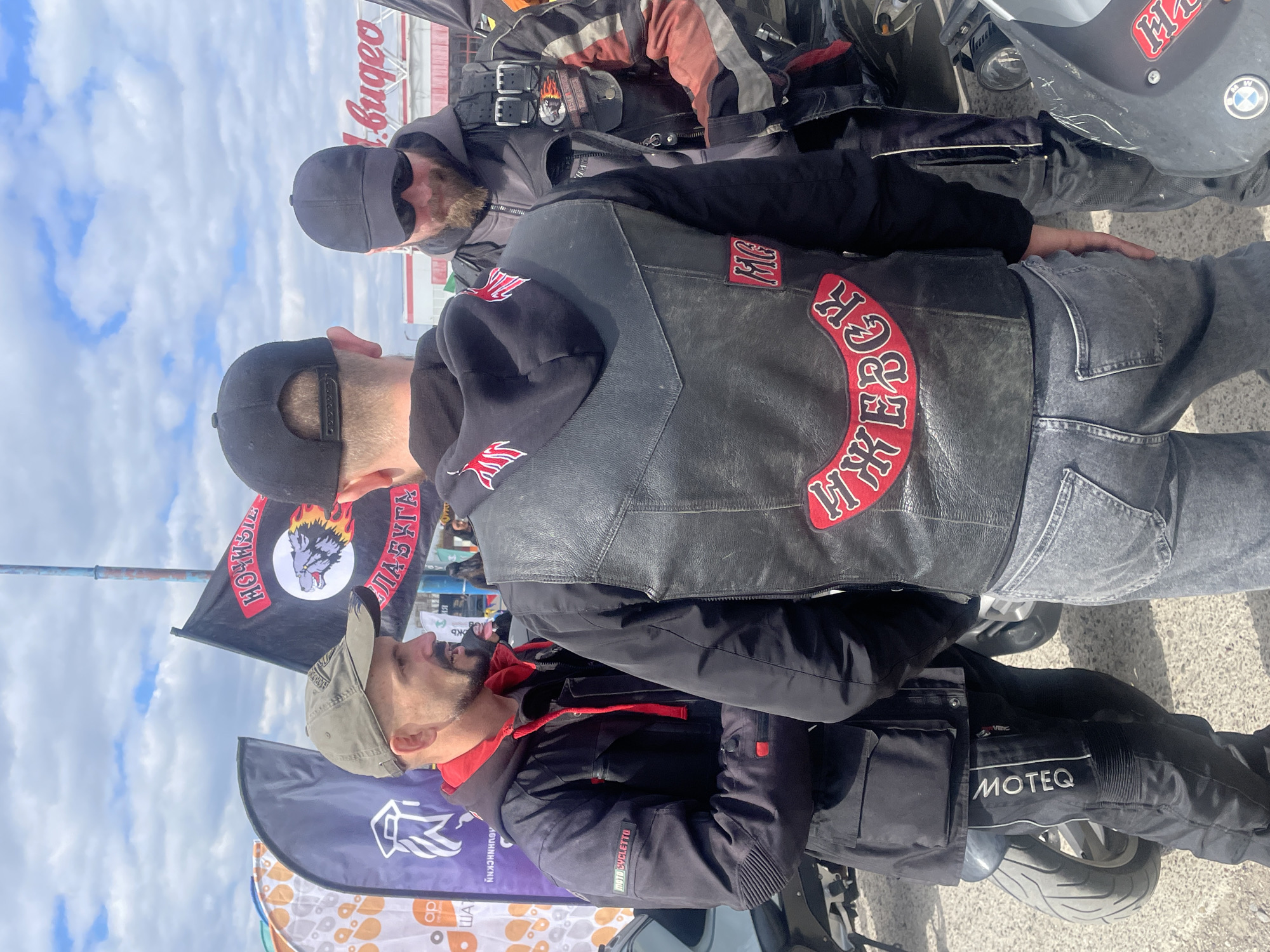 The image shows three men in a public space, likely in Naberezhnye Chelny, Russia, based on the flags of the motorcycle club "Night Wolves" visible in the frame. The men are dressed in motorcycle attire; jackets with motorcycle club patches on them. One man is wearing a black leather vest with a patch with Cyrillic letters that appear to spell out "Night Wolves". The men are near motorcycles, and one man is sitting on the motorcycle. The environment includes other flags and street signs. It is daytime and the weather appears cloudy.