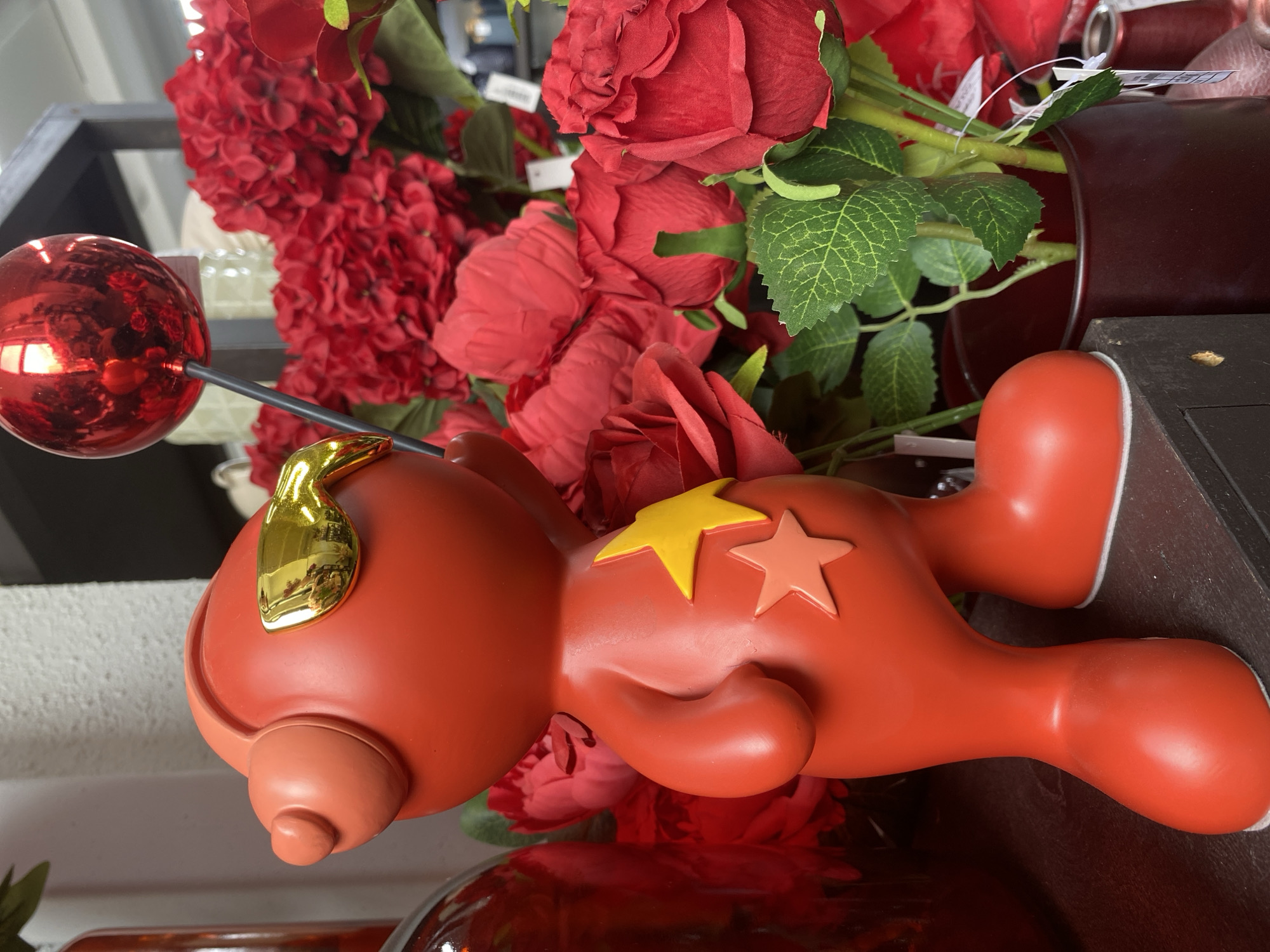 The image features a red, cartoonish figure lying on its side amidst a vibrant display of red roses and hydrangeas. The figure has a glossy red ball attached to a stick protruding from its head, and a shiny golden shape resembling a teardrop near the top of its head. Two star shapes, one yellow and one orange, are positioned on its side. The flowers are arranged in a dark-colored vase or container, with a dark brown or black shelf visible beneath.
The location cannot be determined from the provided information alone.