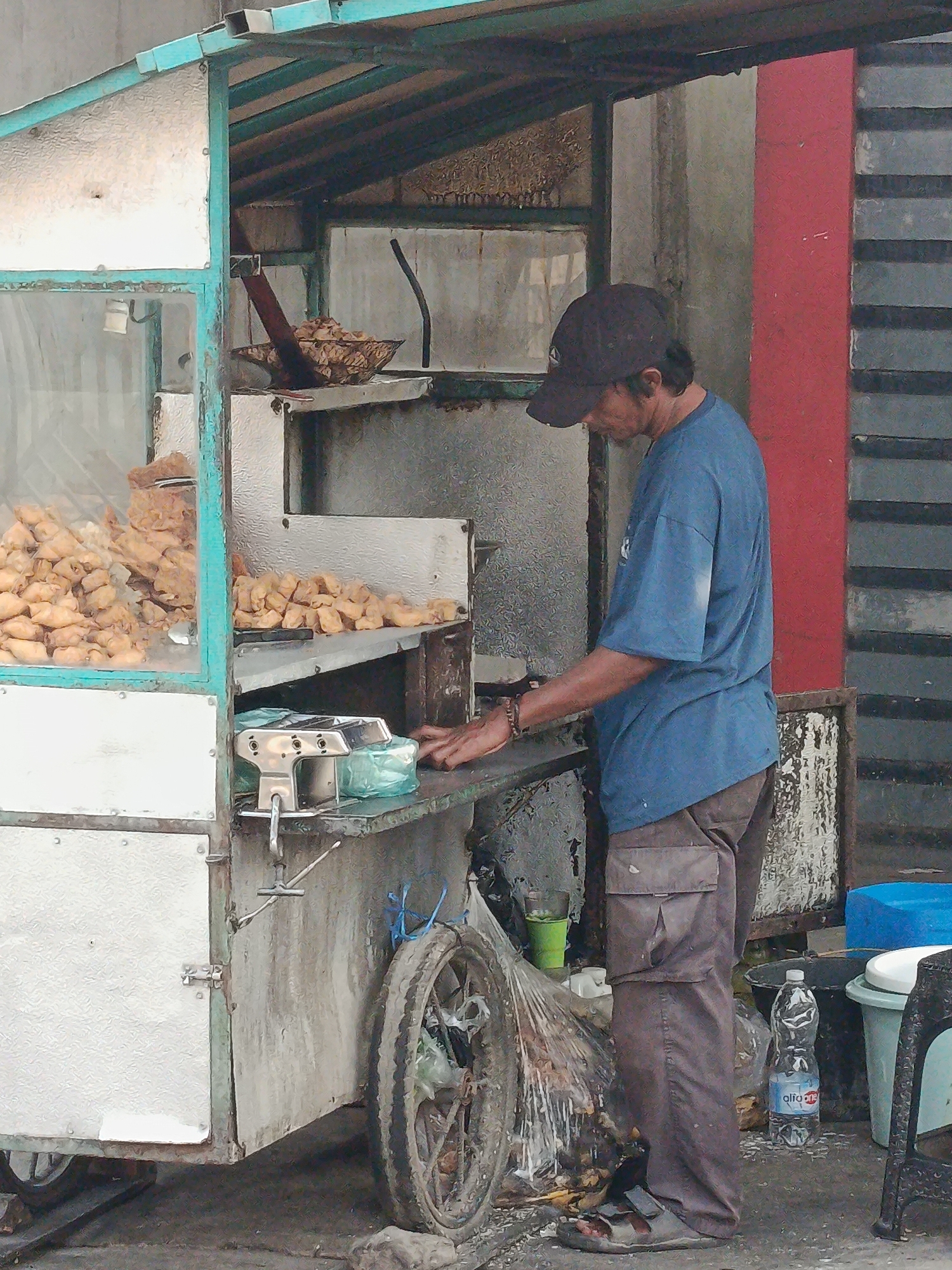 A man, wearing a blue t-shirt, brown cargo pants, and a dark baseball cap, stands behind a food cart in Palembang, Indonesia. The cart is constructed of metal and glass, with shelves holding numerous fried snacks. A scale is visible on the counter, and a bottle of water is on the ground nearby. The man appears to be engaged in preparing or serving food. The environment suggests an outdoor urban setting.