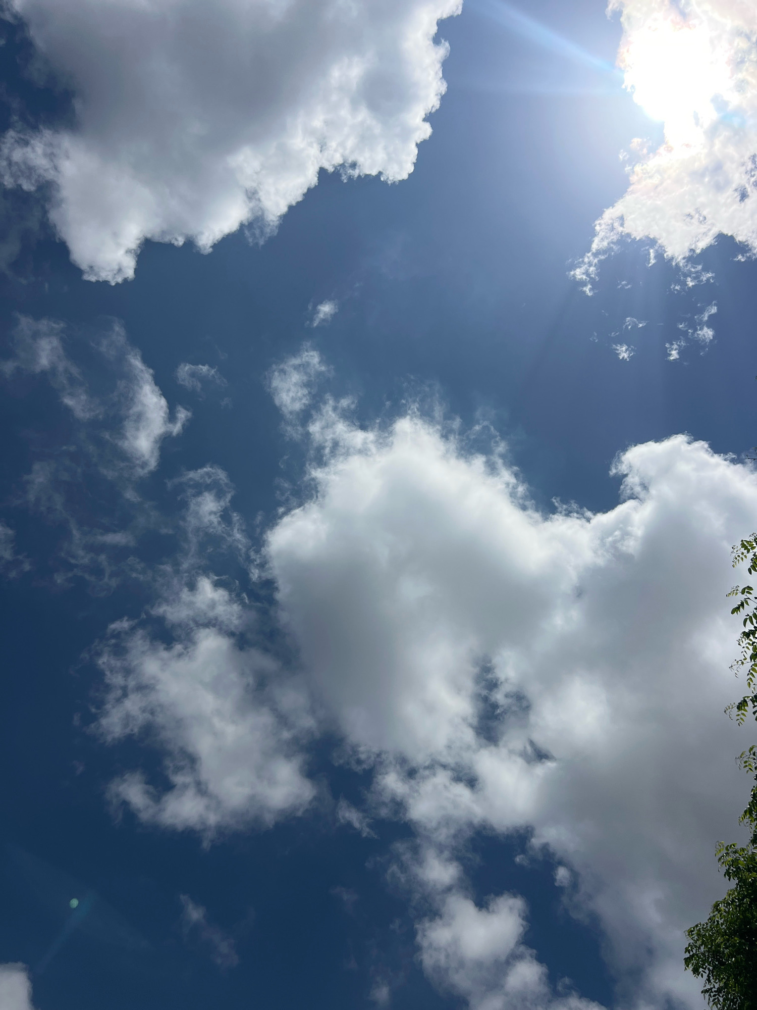 The image shows a bright blue sky with scattered white clouds. The sun is shining brightly, with a lens flare visible in the upper right corner. A few leaves from a tree are visible in the lower right corner. The image appears to have been taken on a clear day in Denpasar, Indonesia. There are no people or any other objects visible in the image, it is a natural landscape.