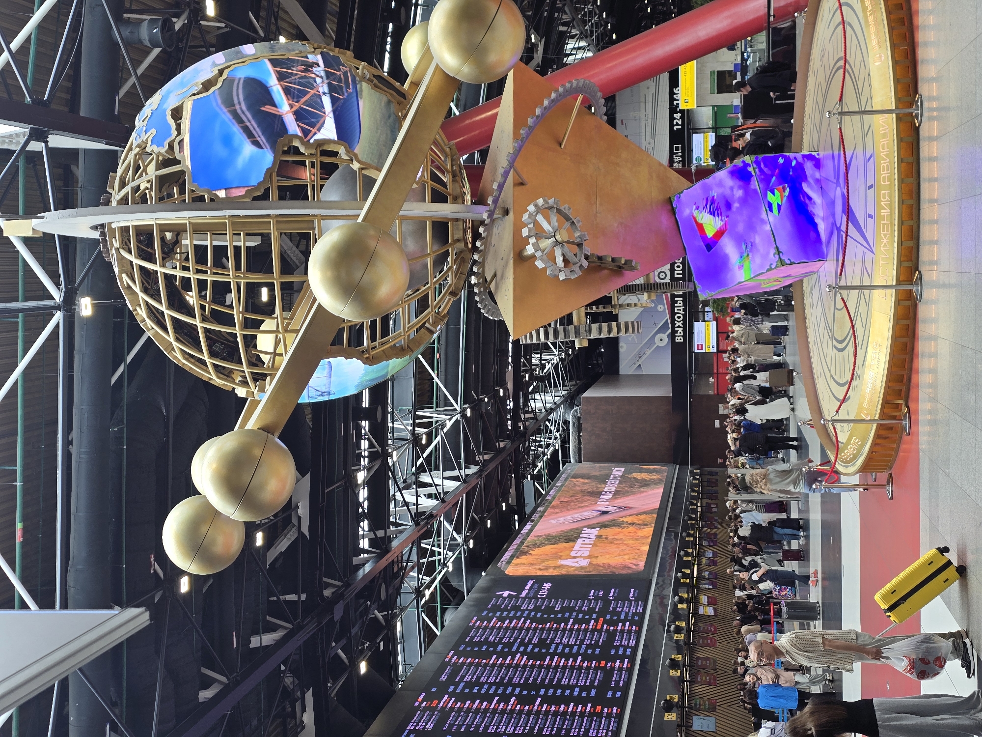 This media file captures an indoor scene within an airport terminal, likely Sheremetyevo Airport in Khimki, Russia, given the visible signage.
The central focus is a large, elaborate kinetic sculpture or art installation hanging from the high ceiling. It features a skeletal golden globe-like structure with integrated screens displaying various dynamic visuals, possibly maps or abstract patterns. Attached to this are golden spherical weights and a triangular wooden structure adorned with decorative gears, suggesting themes of mechanics, time, and global connection. A smaller cubic screen, also part of the installation, shows colorful graphics.
Below and to the right of the sculpture, numerous people are visible, mostly adults. They appear to be travelers, some with luggage, standing in queues or waiting areas. A bright yellow suitcase stands prominently in the foreground.
In the background, a large digital display board shows flight information, including flight numbers, destinations, and statuses, with text predominantly in Russian and English. Above this board, another large screen displays an advertisement or informational graphic with "SITRAK" and "АВСТРАЛИЯ" (Australia) visible.
Further to the right, a circular, ornate platform with a red rope barrier is visible. Text on this platform reads "Шереметьево" (Sheremetyevo) and "ДОСТИЖЕНИЯ АВИАЦИИ" (Achievements of Aviation), confirming the airport setting and the aviation theme of the installation. Other signs, partially visible, include "ВЫХОДЫ К" (Exits to) and gate numbers like "124-146".
The scene is well-lit, characteristic of a modern airport, with exposed structural beams, pipes, and lighting fixtures overhead. The overall atmosphere appears orderly, with people engaged in typical airport activities.