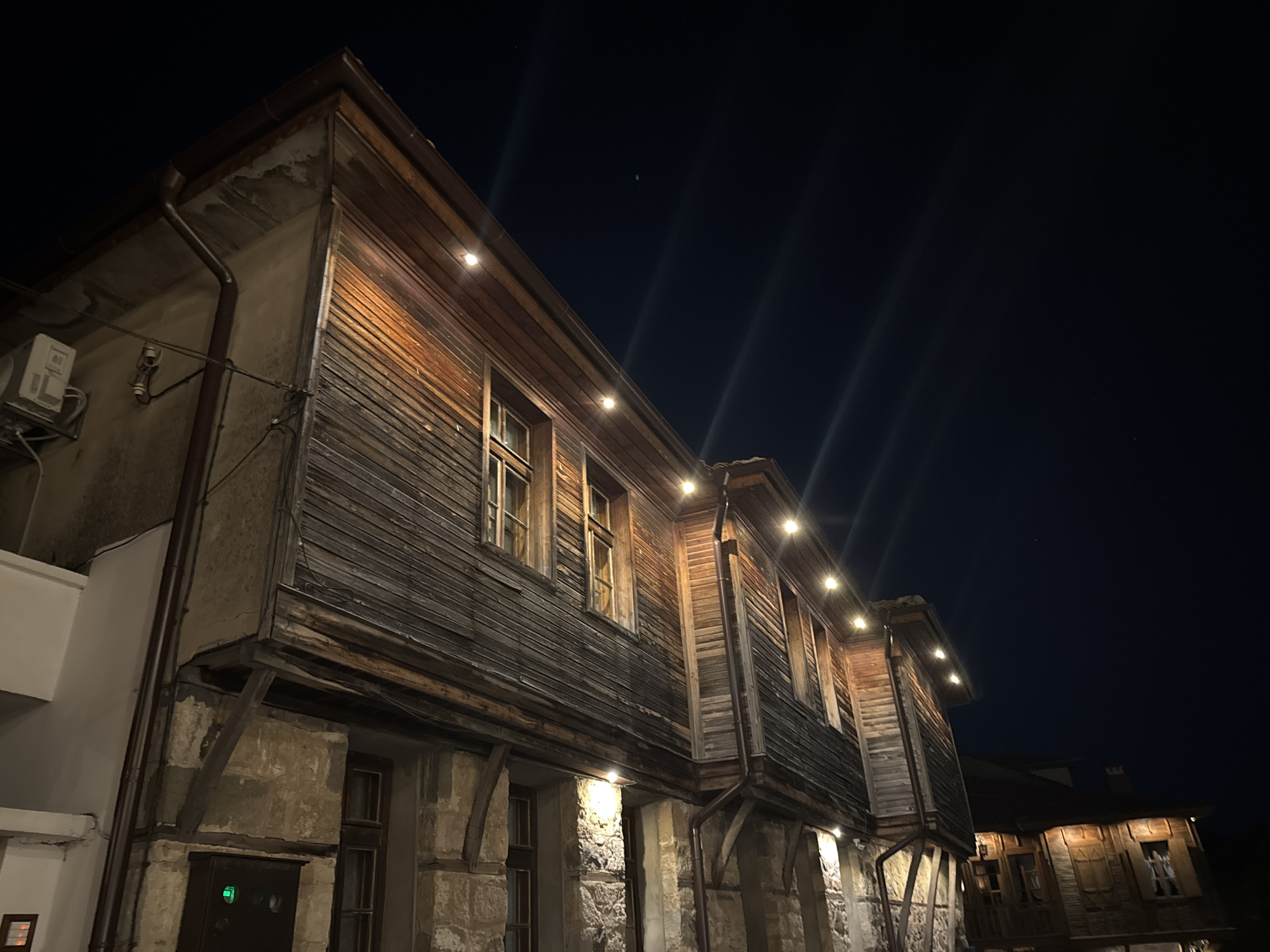 The image shows a view of an old wooden house in the city of Sozopol, Bulgaria, at night. The house features a second floor with horizontal wooden siding and multiple windows. The lower floor is constructed of stone and also has windows. Decorative string lights are mounted along the eaves of the second floor, illuminating the facade. A downspout runs along the side of the building. The sky above is dark, with a few faint lights and streaks that appear to be light trails from the camera or reflections. In the distance, another part of the building is visible, also illuminated by lights. The overall impression is one of historical architecture at night.
