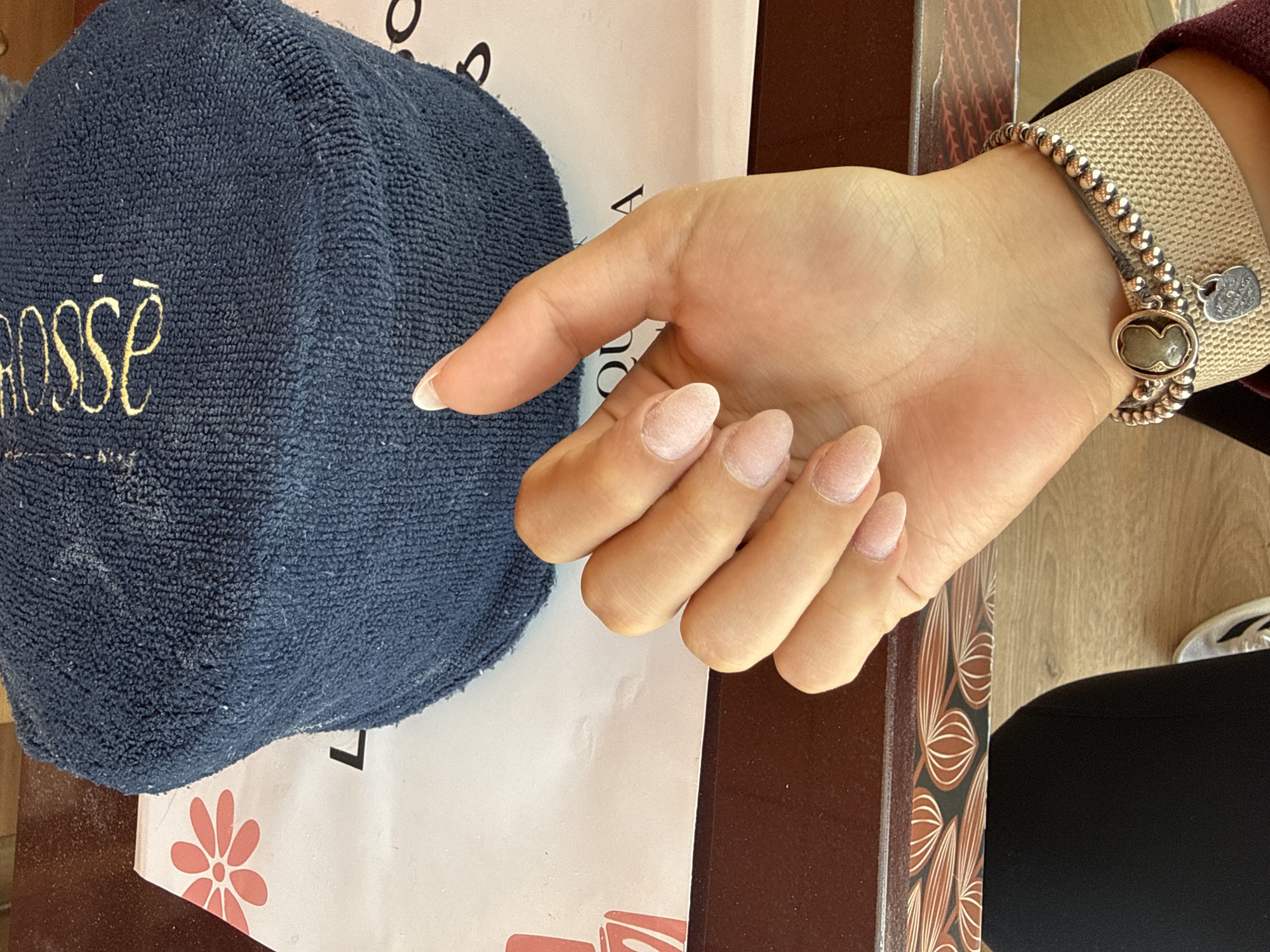 A close-up, high-angle interior photograph depicts a person's left hand and lower arm resting on a dark brown, polished surface within a nail salon located at Rose Quila Salón de Uñas, Av. 16 de Septiembre 3314, El Carmen Huexotitla, Puebla, Mexico. The location is identified by the yellow cursive "Rose" embroidered on a navy blue, textured hand rest, and partially visible black text "QUILA" and a red five-petal floral emblem on a white paper mat underneath.
The subject's left hand exhibits almond-shaped fingernails coated with an unpolished, light pink-toned, textured overlay, indicative of an acrylic or gel application in progress, with the thumb nail showing a more defined white tip. The wrist is adorned with a multi-layered bracelet: a wide, beige woven cuff, two strands of small metallic beads, an oval charm featuring a profile cameo, and a small, silver-toned heart-shaped charm with an embossed design. A dark-colored garment covers the upper leg, and a white shoe with dark accents is partially visible at the bottom. A maroon sleeve is present near the wrist. The navy blue hand rest, positioned behind the hand, is dusted with white particulate matter, consistent with nail filings. The scene indicates an active nail treatment session.