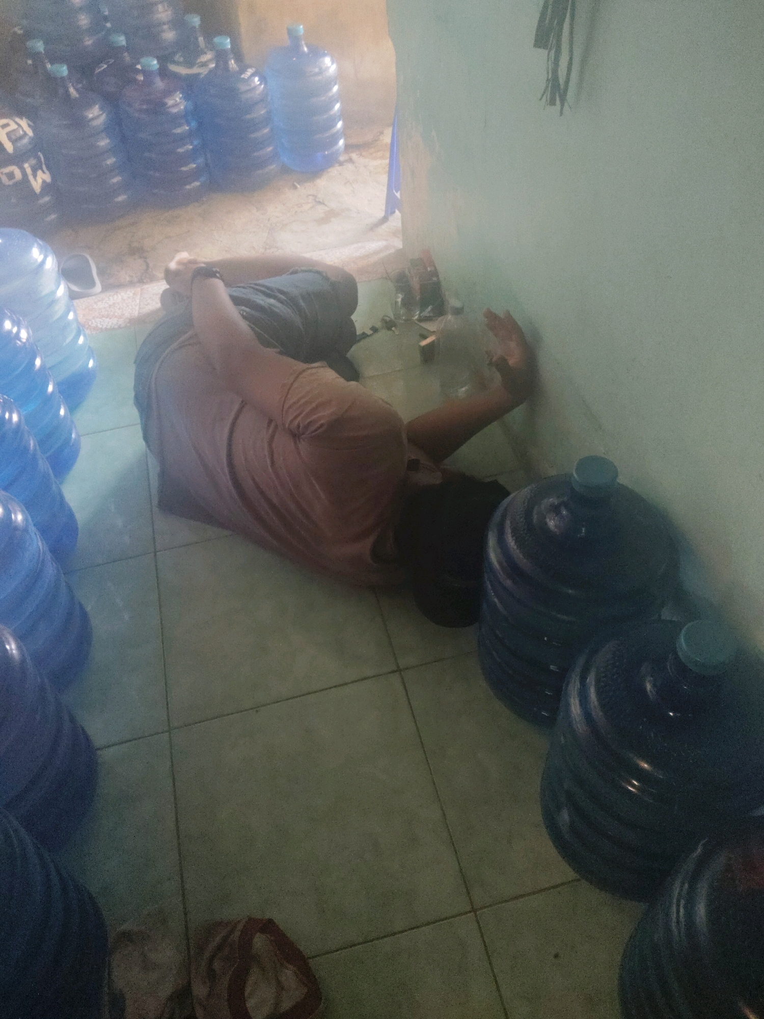A person is lying on the floor next to stacks of large blue water containers in what appears to be a storage room. The person is on their side, with their head resting on their arm and their body stretched out. They are wearing a light brown t-shirt and dark shorts. The floor is tiled, and the walls are a light green color. There are cables hanging from the ceiling in the background. The overall scene suggests a moment of rest or exhaustion within a commercial or industrial setting. The location is specified as a storage area in Subang, Indonesia, potentially a warehouse or distribution center for bottled water.