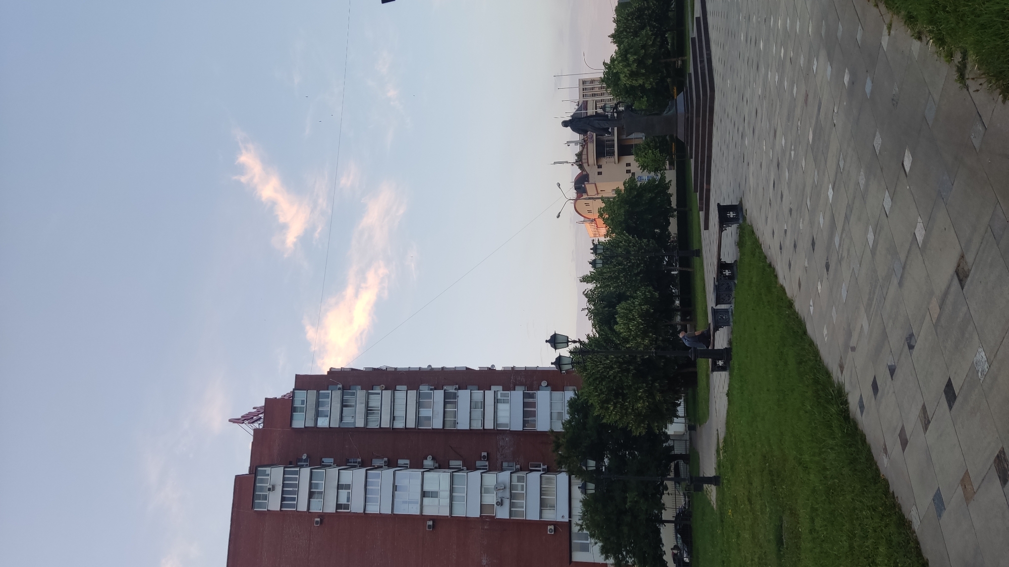 The image shows a view of a city park in Krasnodar, Russia, in the late afternoon or early evening. In the foreground, a paved walkway with a pattern of square and rectangular stones leads into the park. To the left of the walkway is a grassy area with several trees. A park bench is visible in the grassy area with a person sitting on it. Further into the park, there are more trees and lampposts. In the middle ground, a statue of a man on a pedestal stands. Behind the statue, several buildings are visible, including a tall, red brick apartment building with many windows on the left side of the frame. The sky is a pale blue with some wispy clouds, tinged with pink from the setting sun. A thin wire stretches diagonally across the sky.