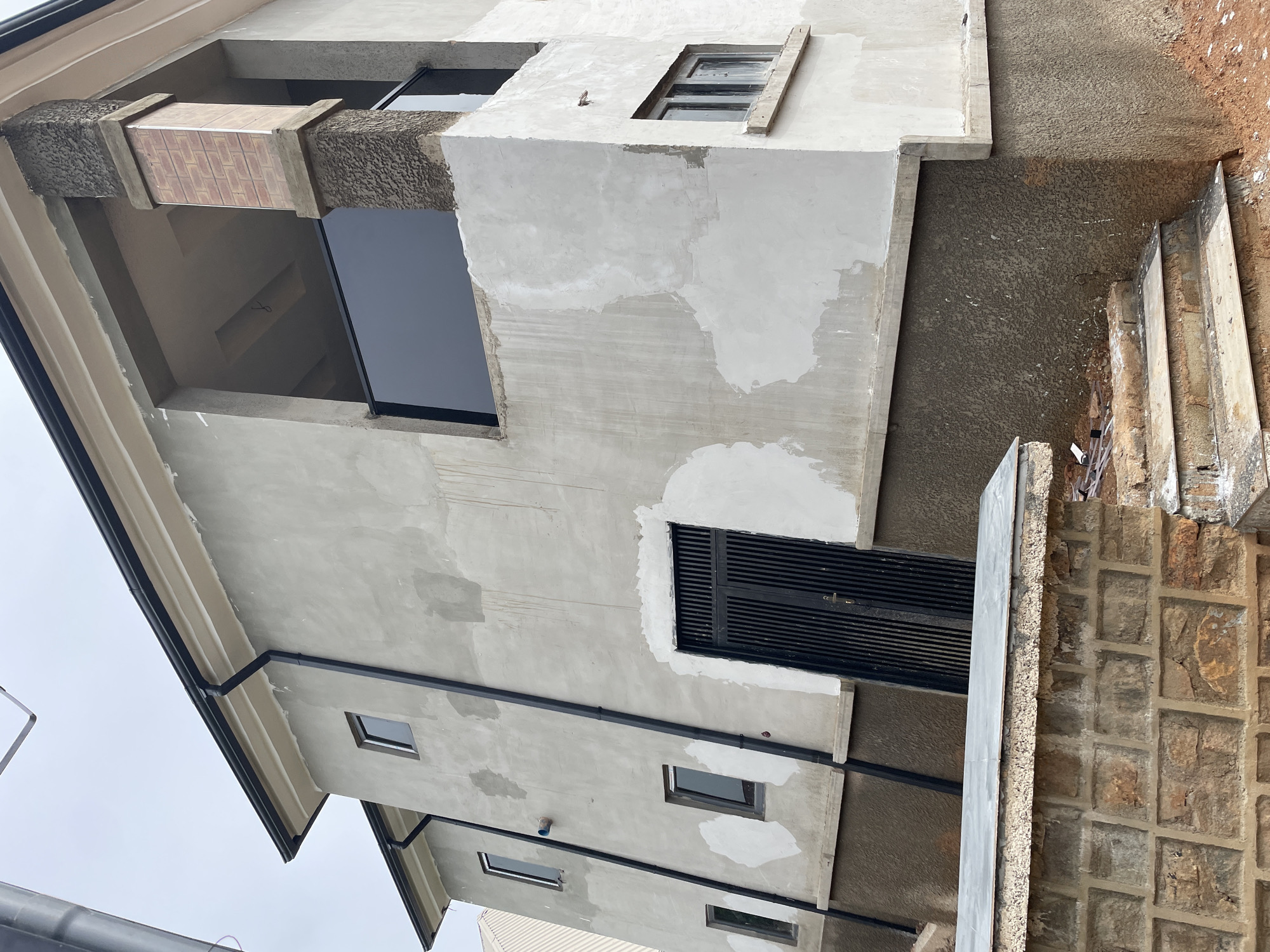 The image depicts the exterior of a multi-story building under construction in Bar Gada, Nigeria. The building has a grey, unfinished concrete facade with some areas showing patches of white plaster. Dark rain gutters run along the roofline and down the side of the building. There are several dark-framed windows, some of which appear to be small and square, while others are rectangular. One prominent feature is a large, dark metal gate.
A balcony area on the upper level is partially visible, supported by a brick and textured concrete pillar. Below the main building, a textured, dark brown base is visible, and a stone wall with a tiled top runs along the lower foreground. Steps made of stone and concrete lead up towards the building. The ground around the building is a mix of red earth and some construction debris. The sky is overcast and pale. There are no people visible in the image.