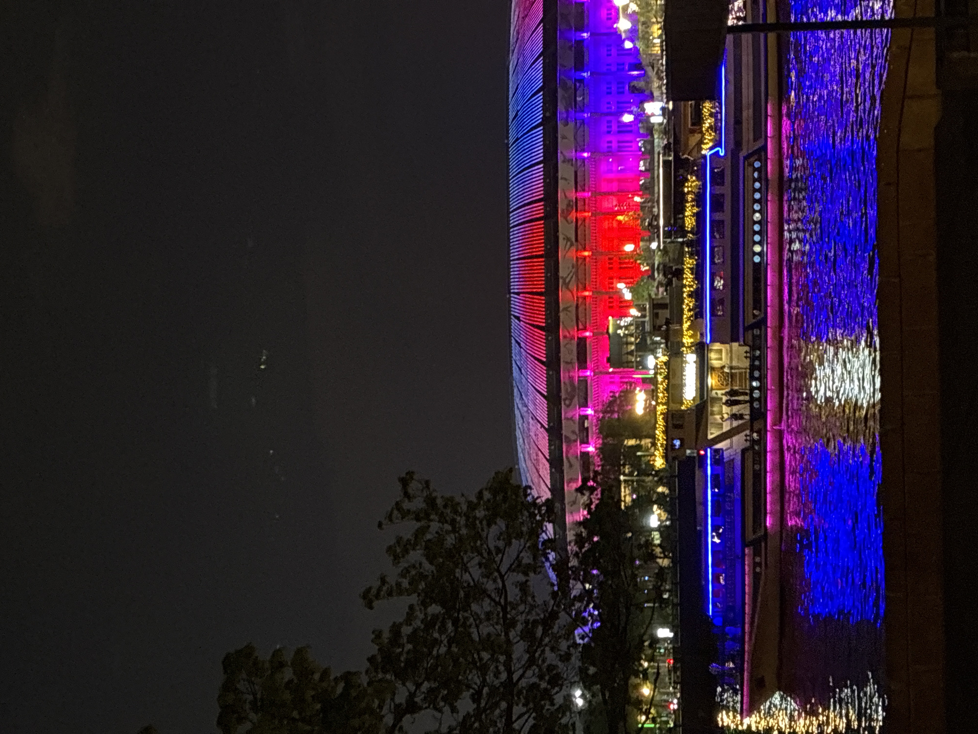 At night, a building in Moscow, Russia, is illuminated with red, white, and blue lights. Reflections of the lights appear in a body of water. Trees and streetlights are visible. A street pole is in the upper right-hand corner.