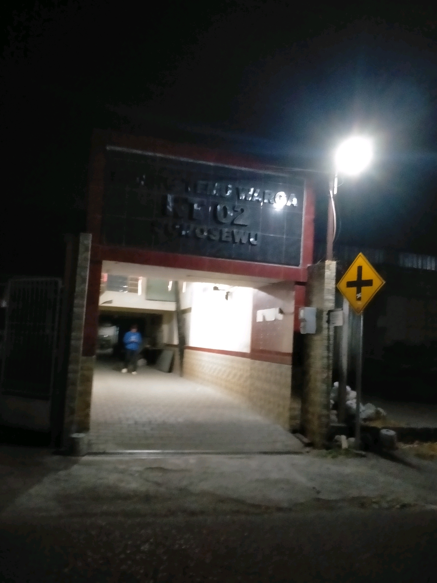 At night, in the city of Kasihan, Indonesia, a building entrance is illuminated by a bright street lamp. The building has a sign with dark lettering on a dark background, partially obscured by the light. The entrance is an archway leading into a well-lit interior with a person visible in blue clothing standing in the doorway. To the right of the entrance, a yellow diamond-shaped sign with a black cross symbol is mounted on a pole. The ground is paved, and there are various objects and debris scattered around the base of the pole and to the right of the entrance. A metal gate is visible to the left of the entrance.