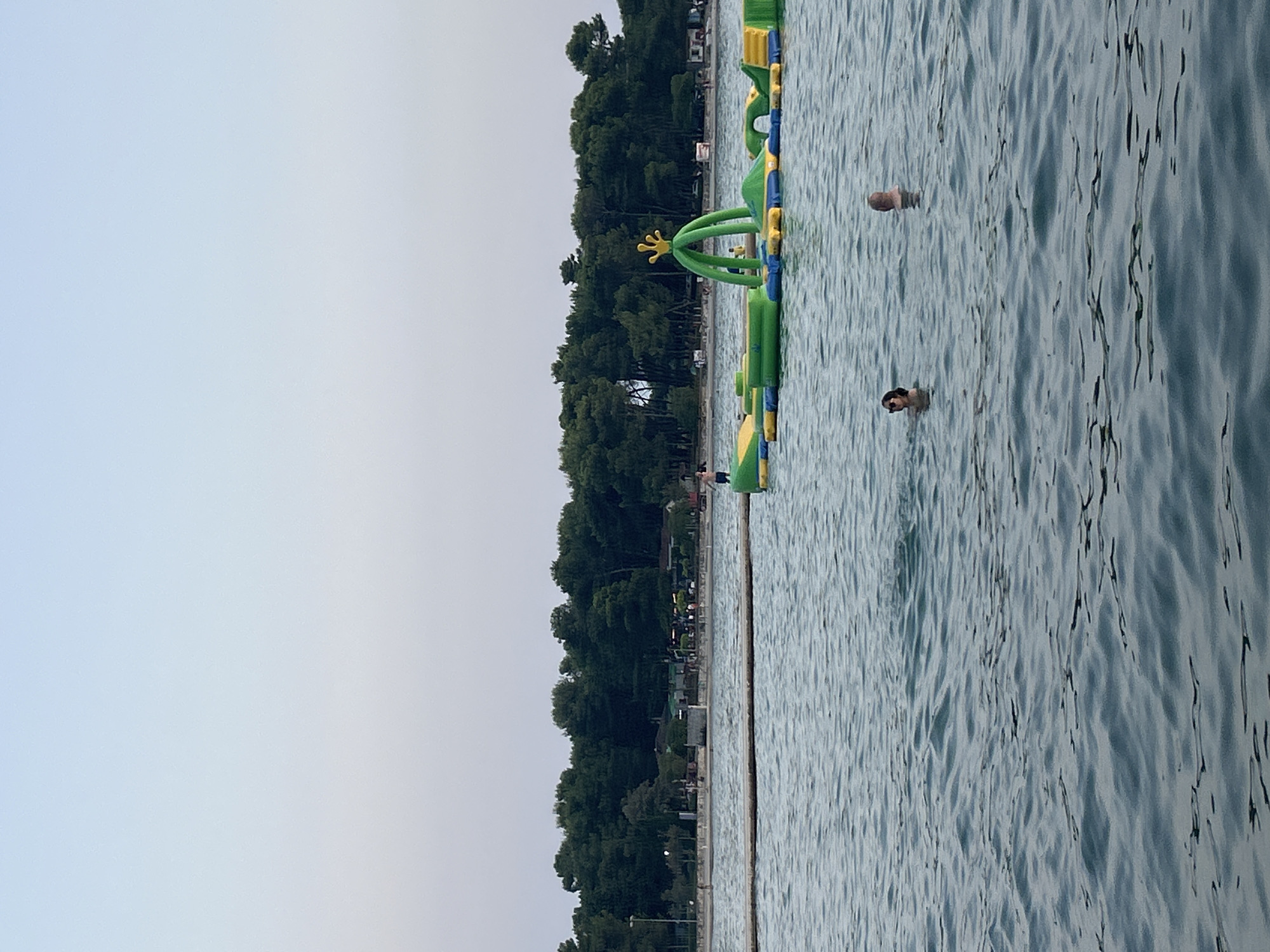 This image depicts a scene at a beach in Katoro, Croatia. The foreground is dominated by the calm, rippling surface of the Adriatic Sea, with two individuals swimming in the water. One swimmer, a woman with dark hair and sunglasses, is partially submerged, facing towards the left. The second swimmer, a man with a shaved head, is also partially submerged, facing towards the right. Further out in the water, an inflatable water park with green and yellow structures, including a large archway with a crown-like top, is visible. A person in blue shorts is standing on one of the inflatable structures. In the background, a long, thin wooden barrier or walkway extends across the water. Beyond the water, a dense line of green trees forms the coastline, behind which a few buildings and structures suggest a resort or residential area. The sky above is a clear, pale blue, indicating it is either early morning or late evening.