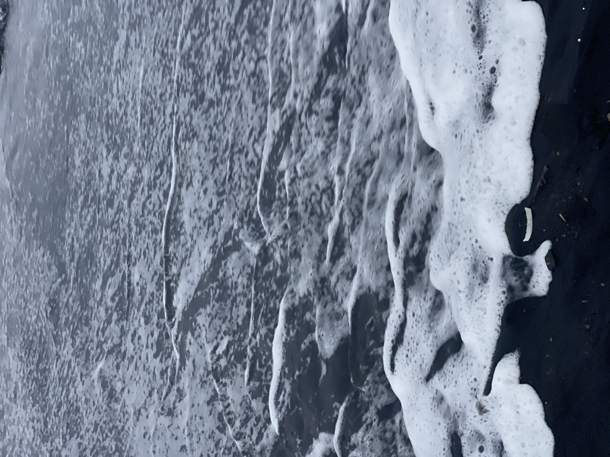 The image shows a close-up view of black sand beach with waves washing ashore. The water is dark and appears to be slightly choppy, with white foam created by the breaking waves. The foam is spread across the sand in some areas, and in other areas, it forms distinct wave patterns. The sand is dark and appears to be wet from the tide. The overall scene is monochromatic, with the dark sand and water contrasting with the white foam. There are no people or man-made objects visible in the image. This scene could be any black sand beach. There is no information available about the specific location in Kediri, Indonesia.