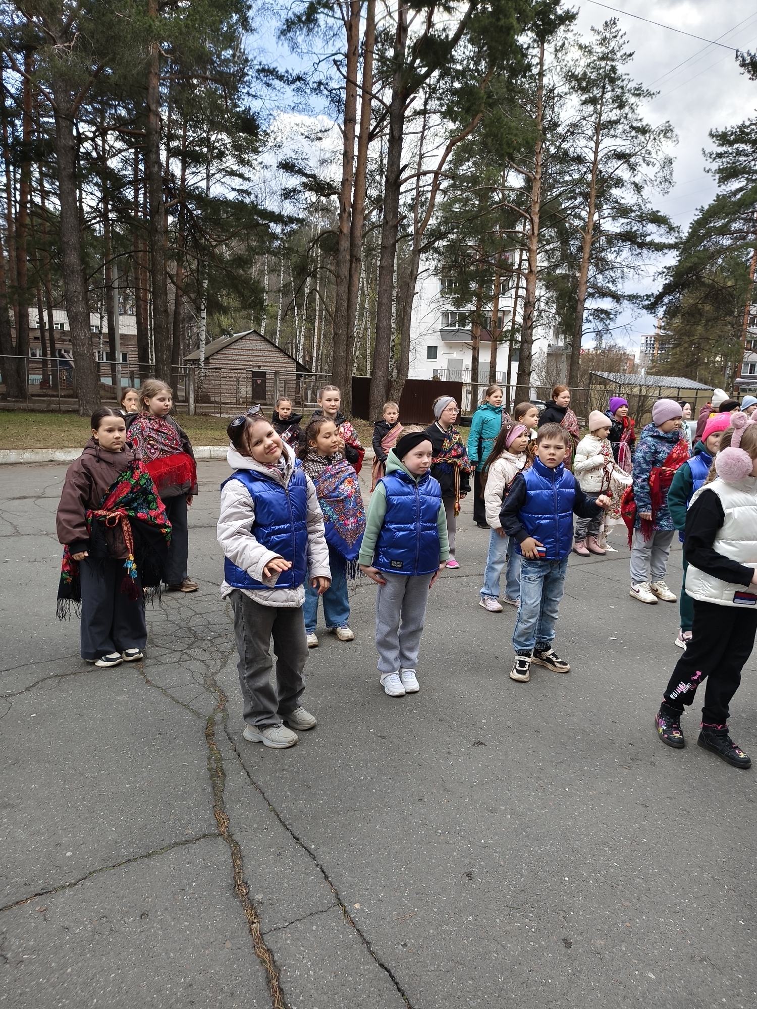 A group of approximately twenty children, boys and girls of varying ages, are gathered outdoors on a paved asphalt surface. Many of the girls are wearing traditional Russian-style patterned shawls (likely Pavloposadskiye platki) draped over their shoulders or head, some in vibrant colors like red and blue. Other children are dressed in modern casual attire, including puffer vests (several in blue), jeans, track pants, and sneakers. One girl in the foreground wears a light gray puffer vest over a white top, with sunglasses pushed up onto her head, and is smiling broadly towards the left. Two boys in the mid-ground also wear blue puffer vests, one of whom has a small patch resembling the Russian flag on his chest.
The children appear to be standing in rows or a formation, suggesting they are participating in an organized event, possibly a cultural performance or gathering. Their expressions range from attentive to smiling.
The setting is an outdoor area, possibly a schoolyard or a quiet street, with tall pine trees dominating the background. Beyond the trees, various buildings are visible, including older wooden structures and more modern multi-story buildings. The asphalt ground where the children stand is visibly cracked in several places, with some weeds growing through. The sky is overcast with patches of lighter clouds, indicating daytime. The children's outerwear suggests cool weather.
Visible text includes the word "EDITION" on the black pants of a child on the far right. Small rectangular patches are visible on the blue puffer vests of two boys, one of which appears to be a Russian flag.