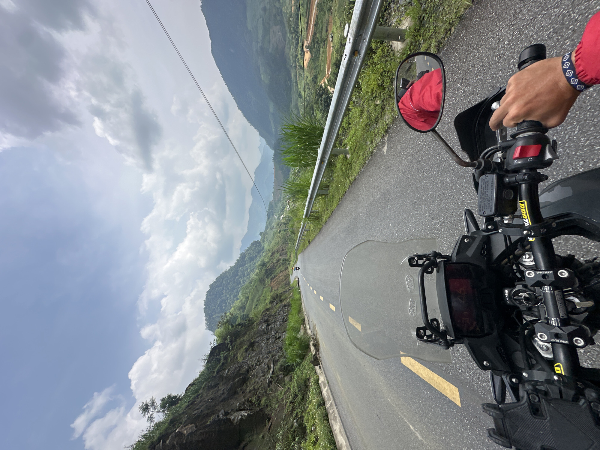 The image presents a first-person perspective from a motorcycle rider on a mountain road. The rider's right hand, wearing a bracelet with a blue and white pattern, grips the throttle, with a rear-view mirror reflecting a portion of the rider's red jacket. The black handlebars, windshield, and dashboard of the motorcycle occupy the lower right foreground.
The asphalt road stretches ahead, marked with a continuous yellow line on the left and broken yellow lines towards the center, leading into a distant curve. A metal guardrail runs along the right side of the road, bordered by lush green grass and dense vegetation on a steep, rocky mountainside. In the midground and background, multiple layers of green, forested mountains rise under a bright, partly cloudy sky, with some power lines visible overhead. The scene suggests a journey through a rugged, natural landscape.
The setting is a winding mountain pass, consistent with the provided context of Minh Sơn Commune, Vietnam. It is daytime, with bright natural lighting. Visible text on the motorcycle includes "SPIRIT BEAST" on the handlebar clamps, and "FRON AS ONLY DOT BRAKE FLUID", "FILLER CAD CONTAINER CLEAN", and "FREIN DOUR" on a brake fluid reservoir or related component.