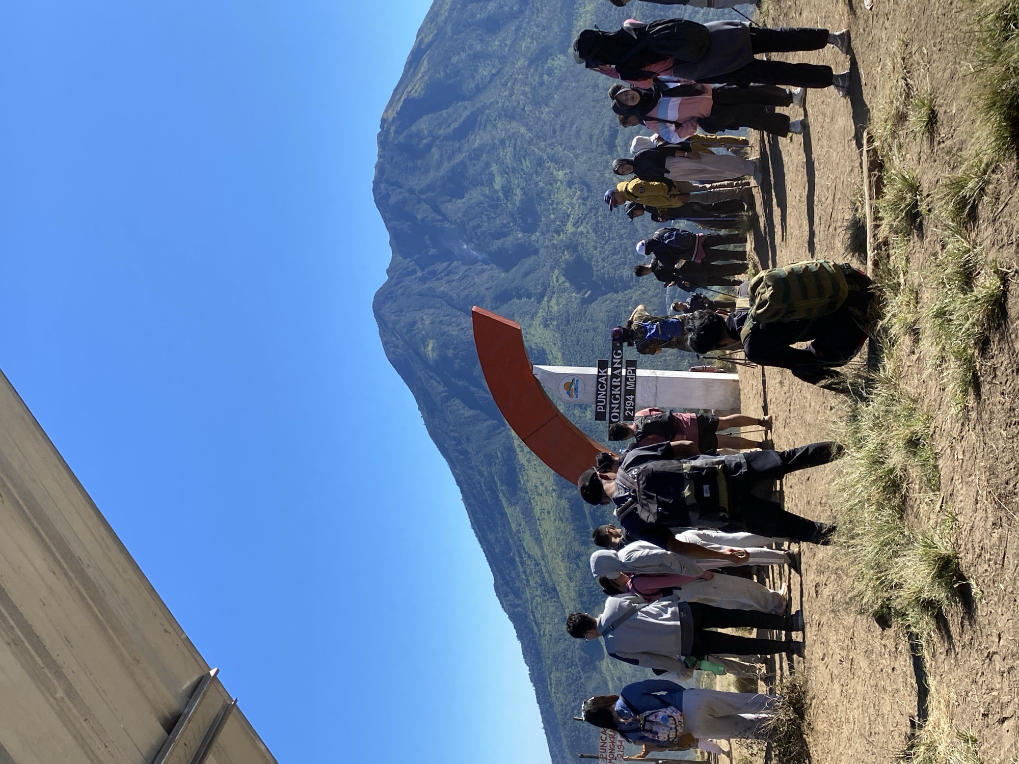 The image depicts a group of people gathered at the summit of Mount Prau, located in the city of Gondosuli, Indonesia. The sky is clear and blue, suggesting good weather. In the foreground, dry grass and dirt cover the ground, with shadows cast by the people. A large mountain, covered in lush green vegetation, dominates the background. A signpost indicating "PUNCAK ONDERKRANG 2194 MdPl" is visible, marking the summit. A large, curved red structure is positioned near the signpost. The people are dressed in casual clothing, many with backpacks, indicating they are hikers. Some are looking towards the mountain, while others are interacting with each other or taking photos. The overall scene conveys a sense of accomplishment and shared experience at a scenic mountain peak.