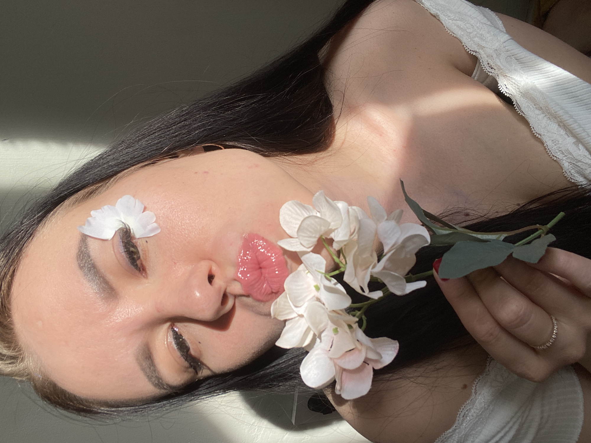 A light-skinned woman with long, dark hair lies down, facing upwards and angled to the left. She wears a white lace-trimmed top. Her eyes are closed. A white flower petal rests on her forehead, and a larger cluster of white and pink flowers is positioned near her mouth. An artificial-looking pair of glossy red lips is photoshopped over her own mouth. She holds the flower stem in her right hand. Her right hand also features a red fingernail and is adorned with a ring on her right ring finger. The background is indistinct, but appears to be a room with light-colored walls. The location of the image is not discernible.
