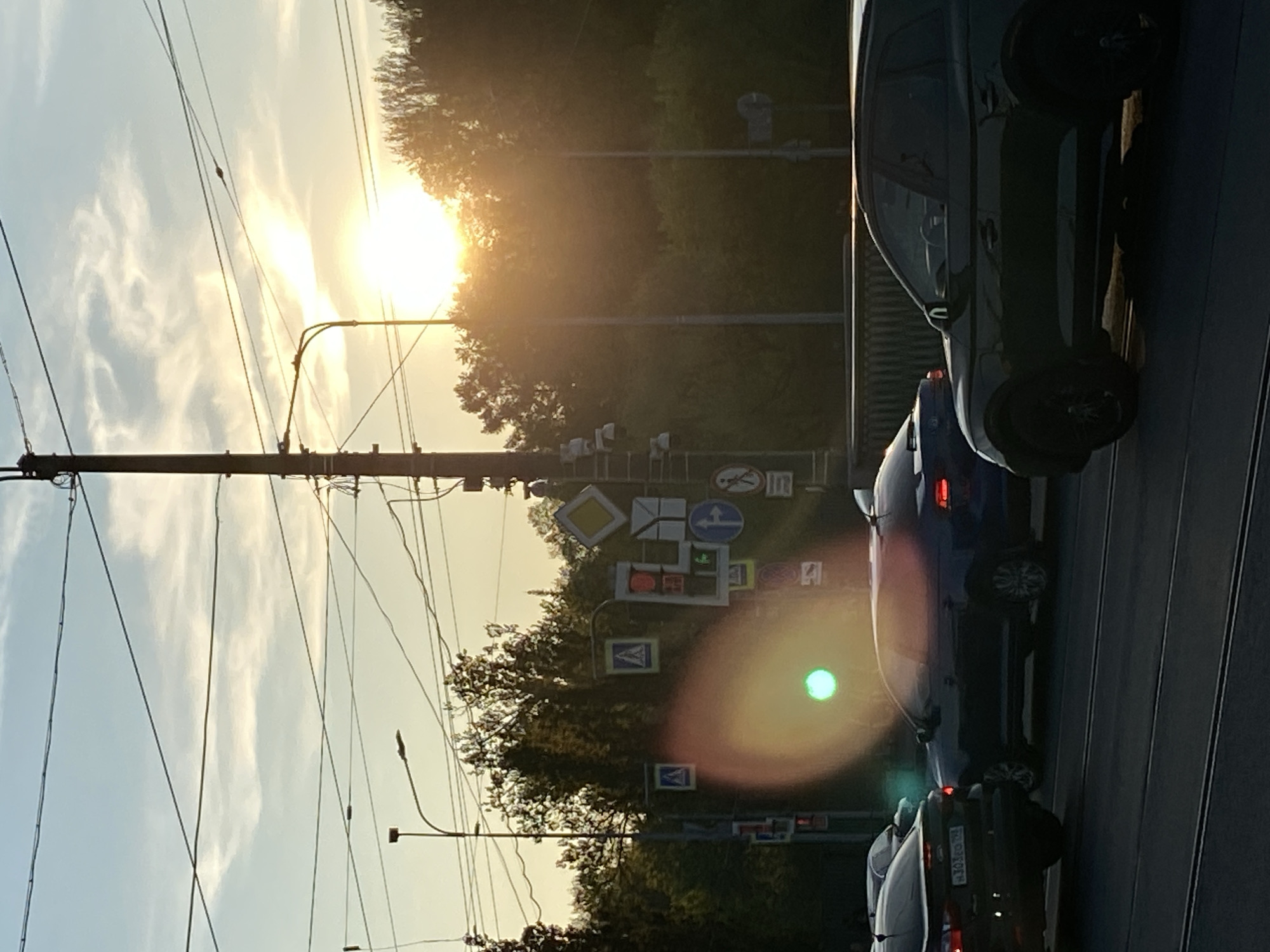 The image depicts a street scene in Saint Petersburg, Russia, at sunset. A power pole with numerous wires is visible on the left. Various road signs are mounted on a vertical structure in the middle ground. Several cars are lined up on the road to the right. The sunlight is filtering through the trees in the background, creating a bright, hazy effect. Tram rails are visible in the lower right.