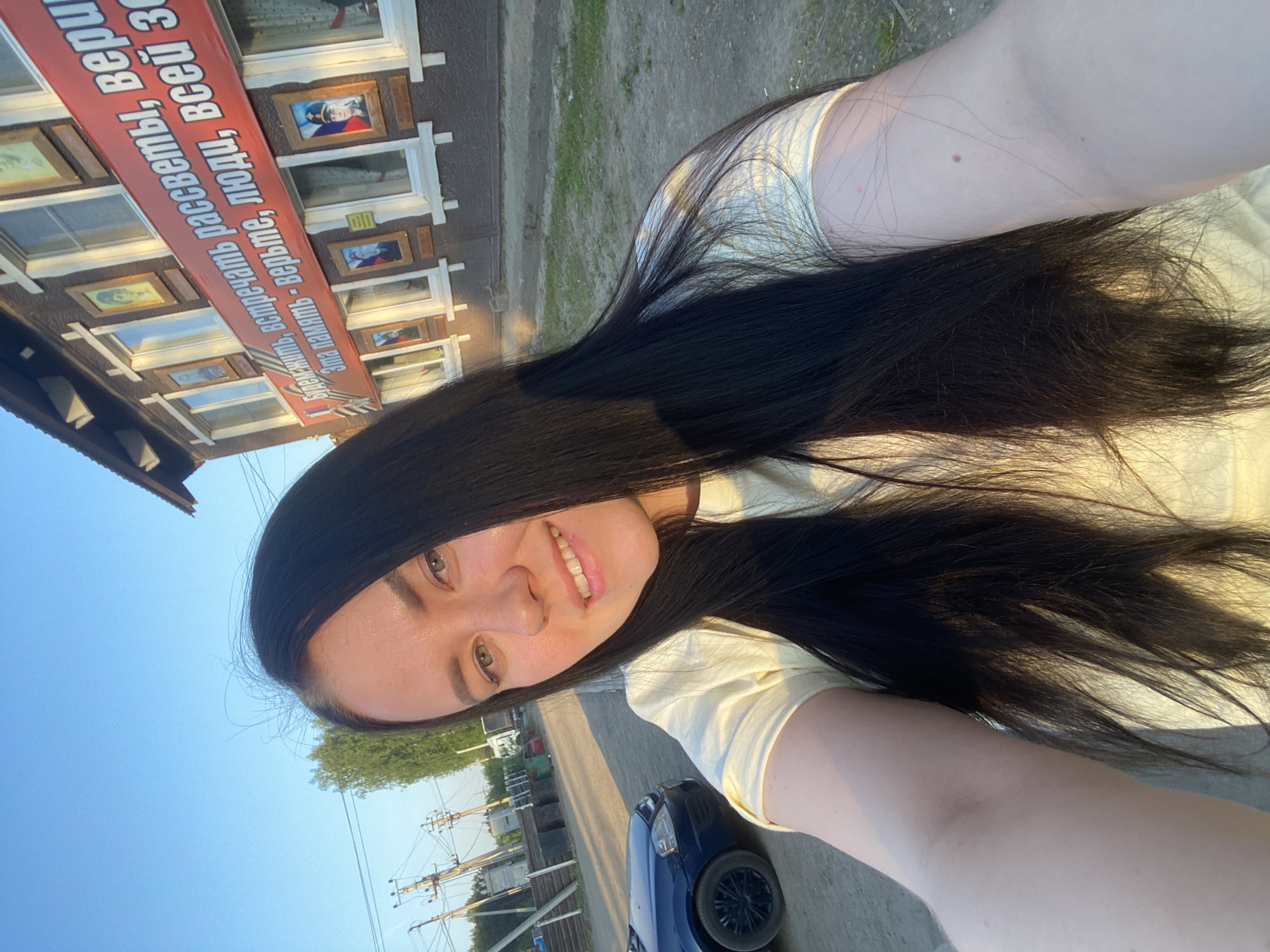 A young woman with dark hair is taking a selfie. She is smiling. Behind her is a building adorned with framed pictures and a sign written in Russian: "Genit, meet the sunrises, trust. This memory - believe, people, of all". A car is parked on the street next to her. Power lines run along the street. The location is in the city of Noviy, Russia.