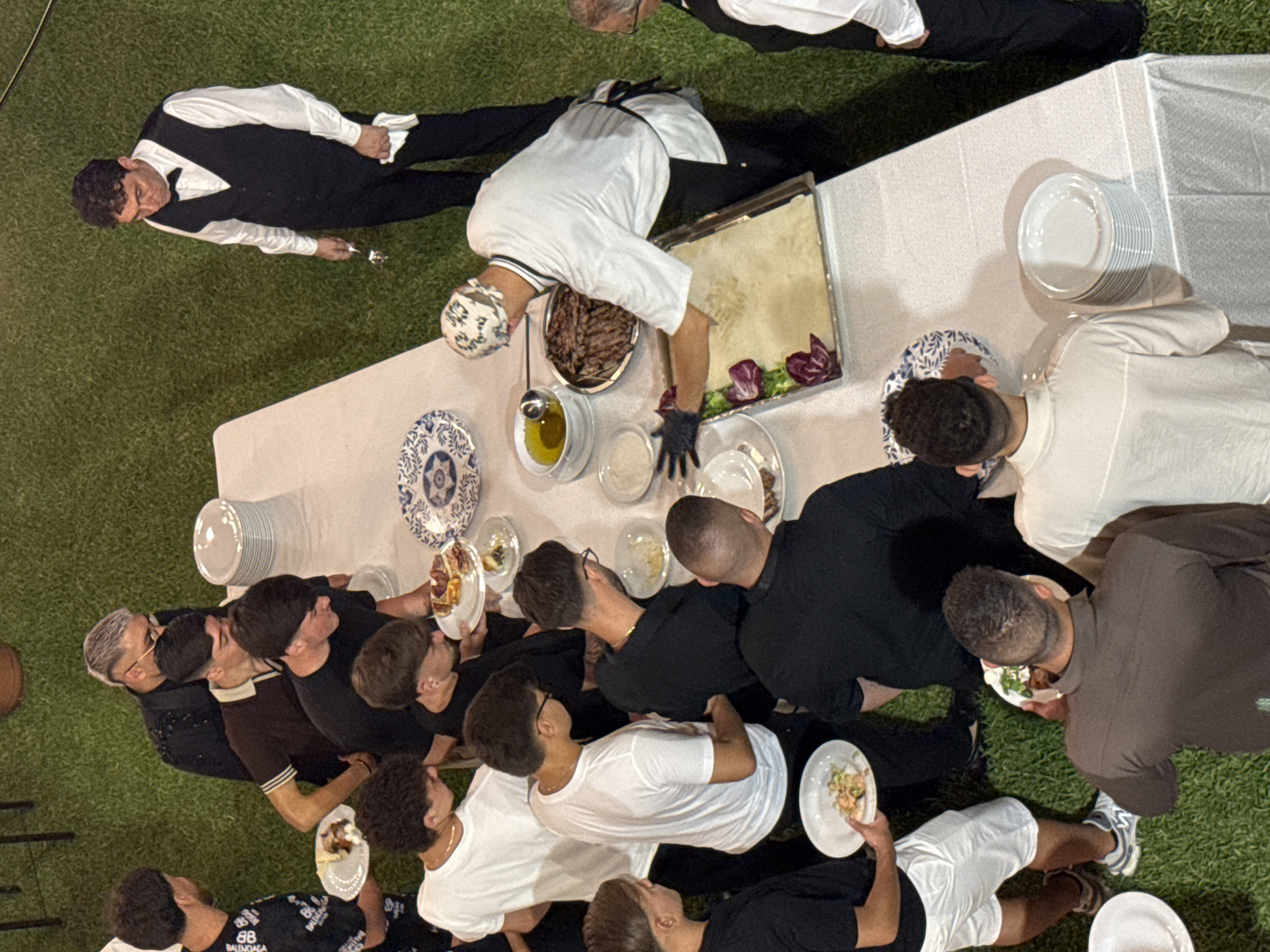 In Isola delle Femmine, Italy, an outdoor reception is taking place on a grassy area. A long table is set up with a white tablecloth, laden with plates of food and stacks of clean plates. Several people, predominantly young men, are gathered around the table, some holding plates with food. A chef in a white uniform and bandana is actively preparing food on a tray, garnishing it with red lettuce. Another man, dressed in a black vest and bow tie, stands nearby, holding a fork and napkin. The overall atmosphere appears to be a casual outdoor event with a buffet-style meal.