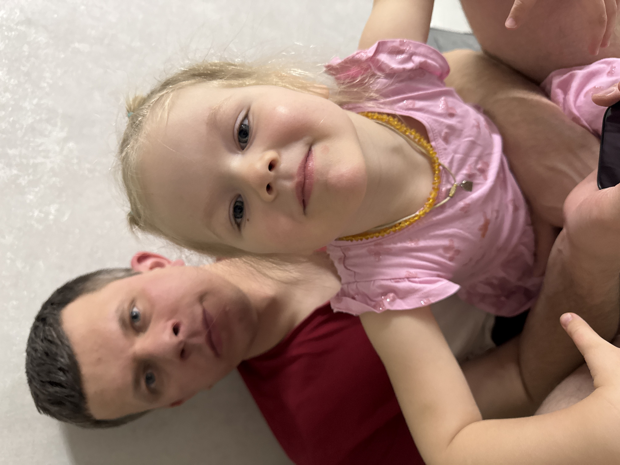 In an indoor setting within the city of Gornyi (Dvurechenskoe S/P), Russia, an adult male and a young female child are depicted. The adult male, with short dark hair and blue eyes, wears a red short-sleeved shirt and is seated in the background, holding the child on his lap. The child, with blonde hair styled in two small pigtails and blue eyes, wears a pink short-sleeved garment with a subtle pattern and an amber beaded necklace with a small metallic charm. She is positioned in the foreground and is in sharper focus than the adult. Both subjects are looking towards the camera. A dark-screened mobile phone is partially visible in the bottom right corner, held by unseen hands. The background consists of a plain, light-colored, textured wall.
