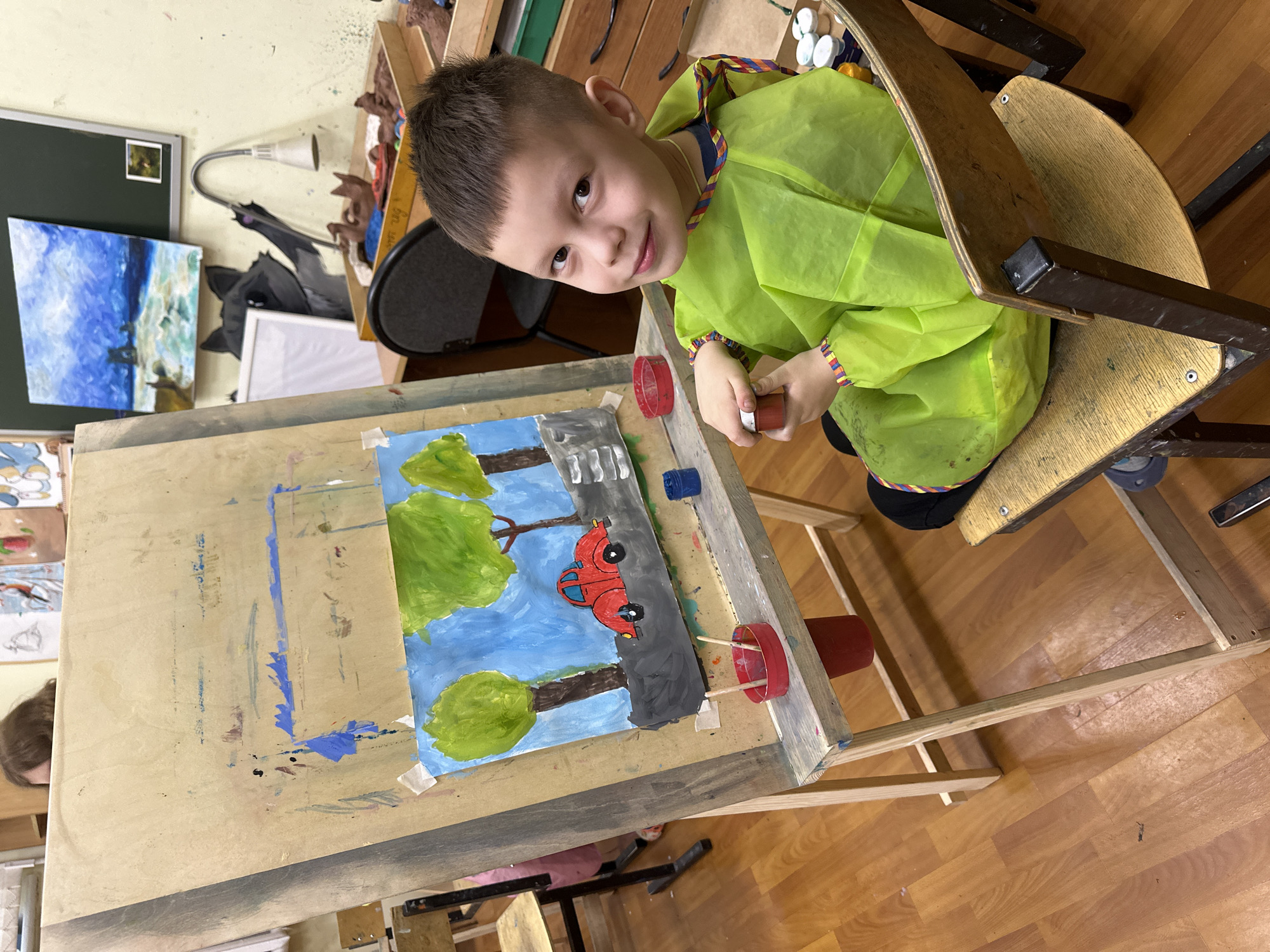 A young boy, likely of elementary school age, is captured in an art classroom or studio in Tomilino, Russia. He is the central subject, positioned in front of a wooden easel, looking directly at the camera with a slight smile. He wears a bright lime green art smock with a multi-colored trim on the collar and cuffs, indicating he is engaged in a painting activity.
On the easel is a colorful painting on paper, depicting a blue sky, two green trees with brown trunks, a grey road with white dashed lines, and a red car. The painting is taped to the wooden surface of the easel, which is splattered with various paint marks. The boy holds a small red paint container in his hand, with a blue container also visible near the painting.
The setting clearly indicates an art space. In the background, shelves display various art creations, including brown sculpted forms and a blue clay object. A dark board, possibly a blackboard, with a small image pinned to it, and a flexible desk lamp are also visible. To the left, another painting with a blue sky and water motif is partially visible. The boy is seated on a wooden chair with a metal frame, and the room has a polished wooden floor. The overall scene suggests a creative and engaging environment for artistic expression. No discernible text is visible within the image.