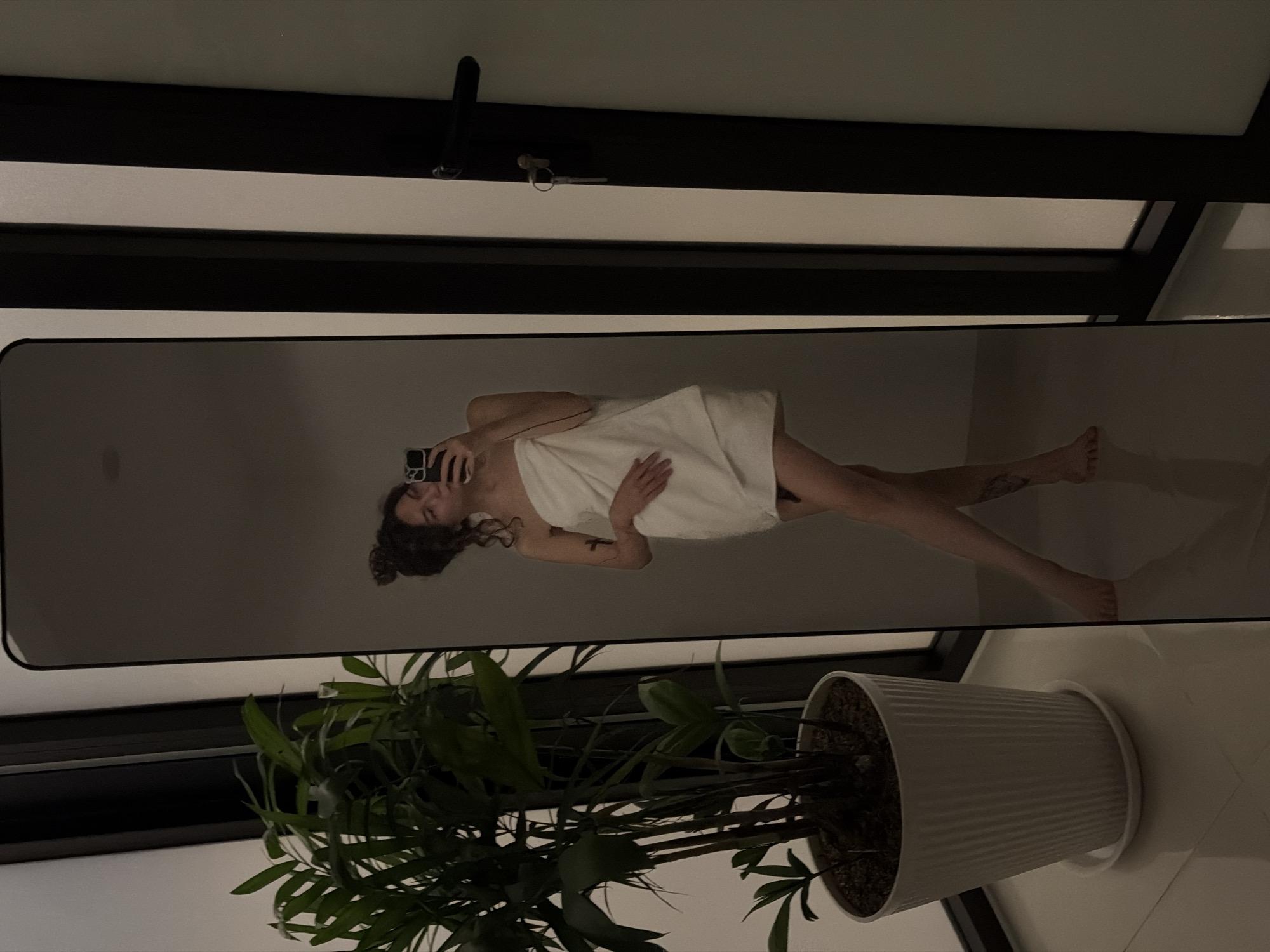 A mirror reflects a woman wrapped in a white towel, holding a phone, presumably taking a photo of herself. She has dark hair and fair skin, and appears to be in an indoor setting. In front of the mirror is a potted plant with green leaves. The background shows a neutral-colored wall and what appears to be part of a window frame. There is no information in the image that indicates the city or location of Đà Nẵng, Vietnam.